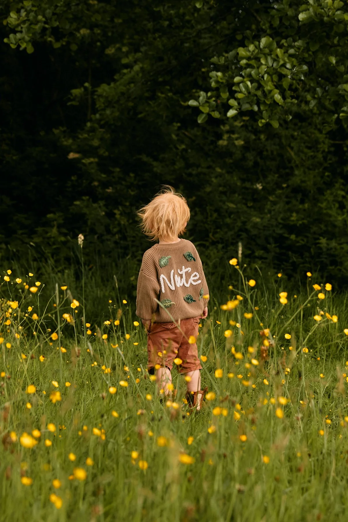 A young child with blonde hair standing in a field of yellow flowers, wearing a brown sweater with the word 'Note' and green leaf decorations, and shorts, with a dark green forest in the background.