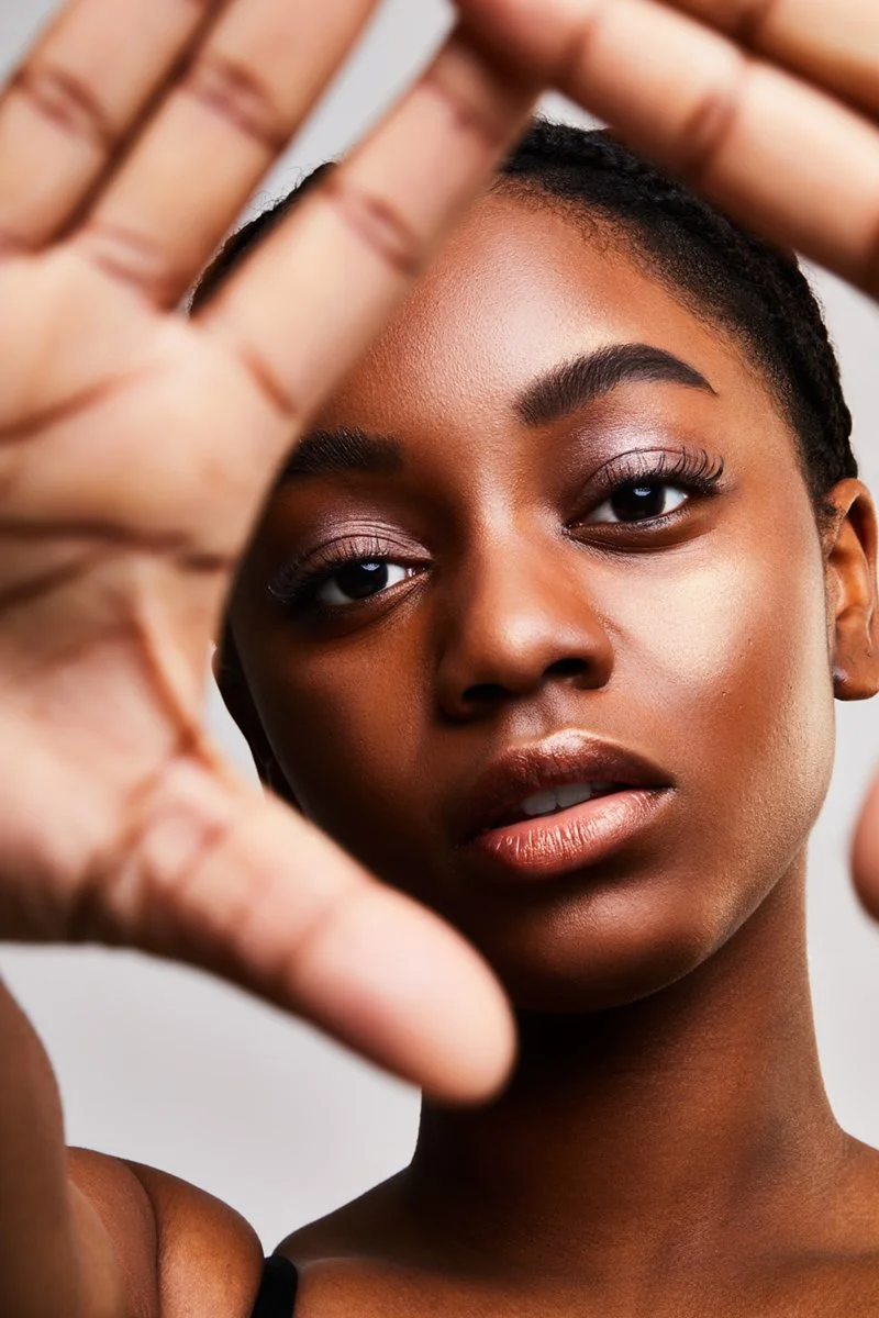 A close-up of a woman with dark skin, short hair, and makeup, holding her hand in front of her face.