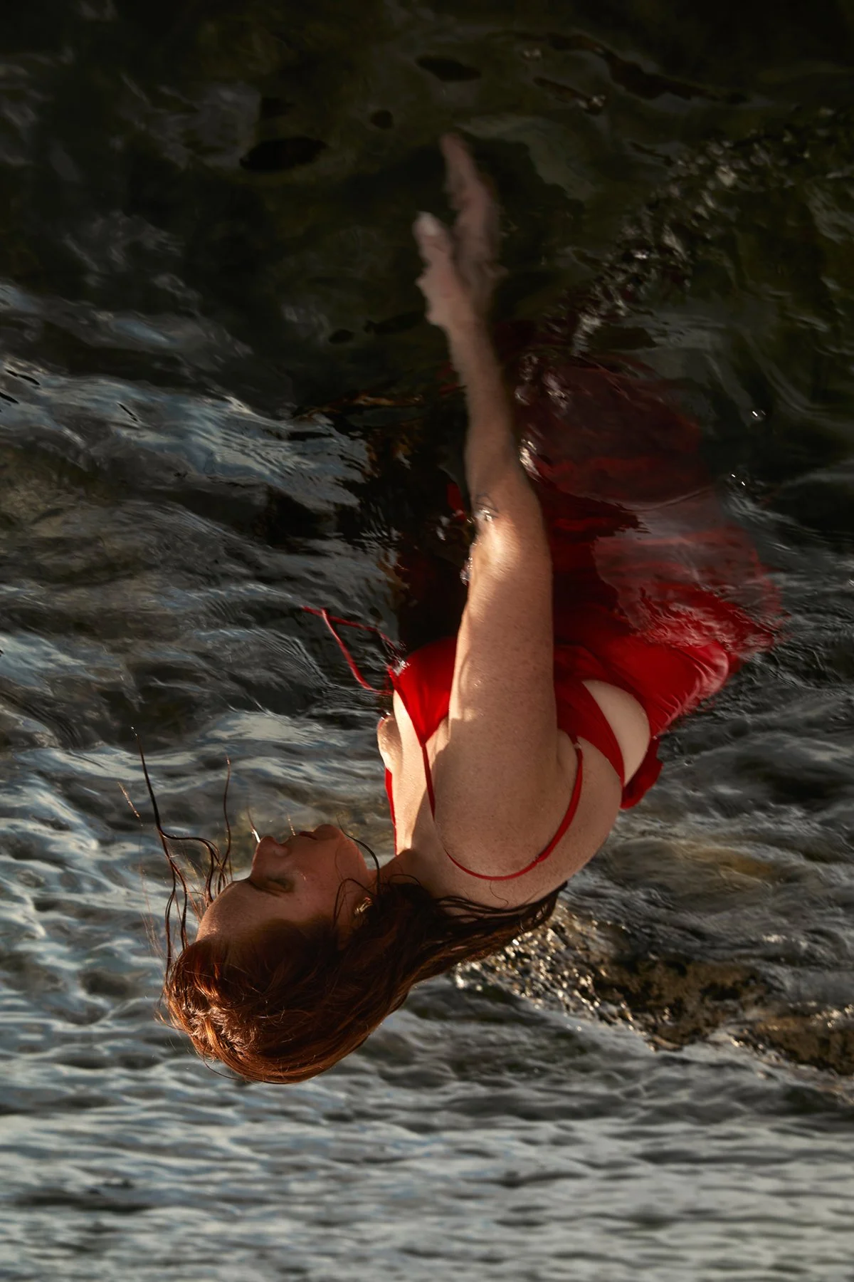 A woman in a red dress floating in a body of water, with her head tilted back and wearing earrings.