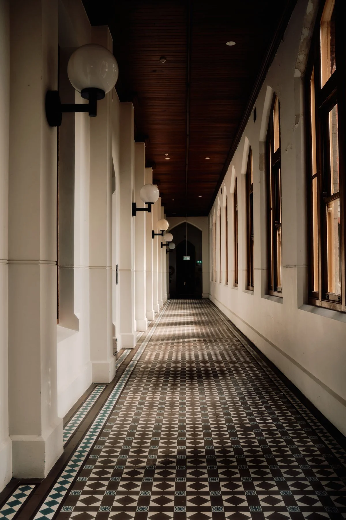 Interior of a hallway with patterned tile floor, large windows, and wall-mounted globe lights.