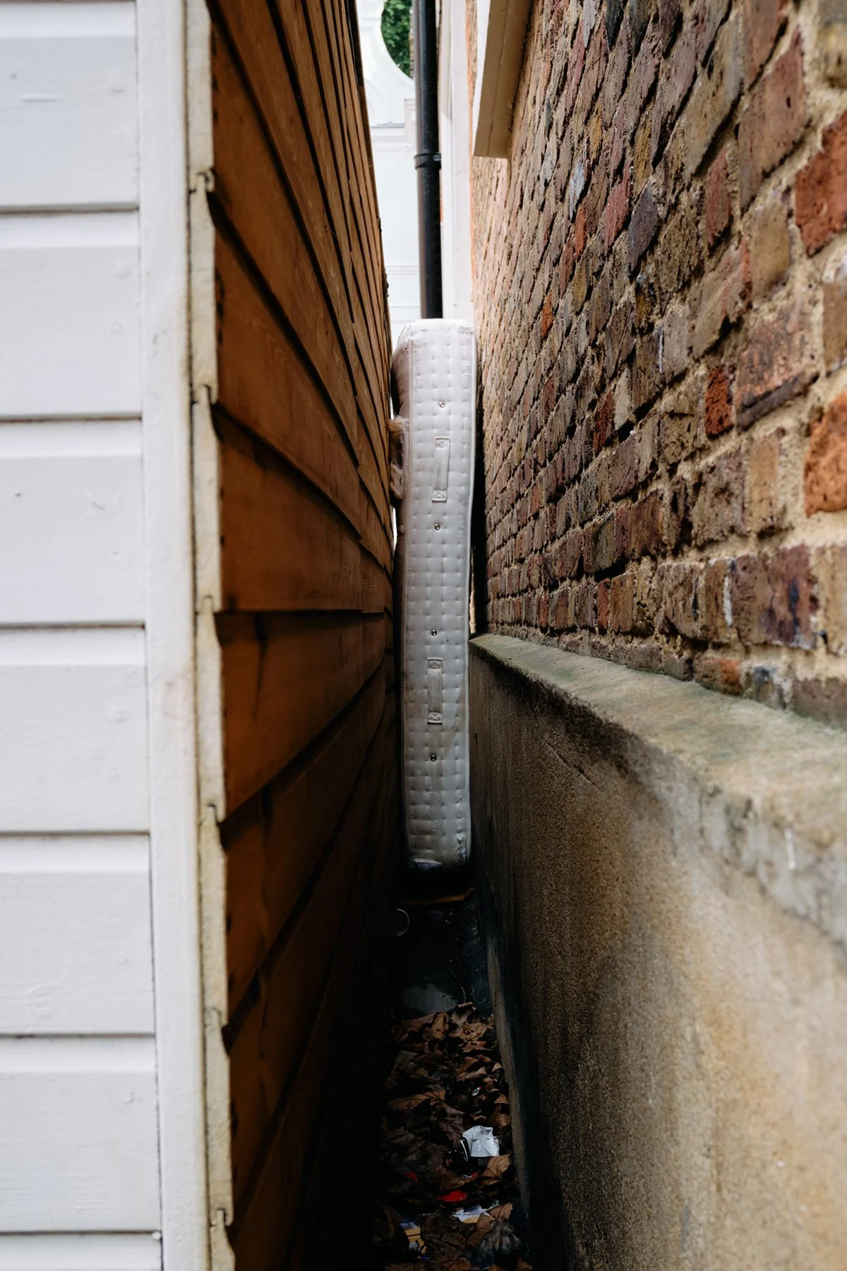 Narrow alley between a white house with horizontal siding and a brick wall, with a stacked mattress leaning vertically against the brick wall, and leaves and trash on the ground.