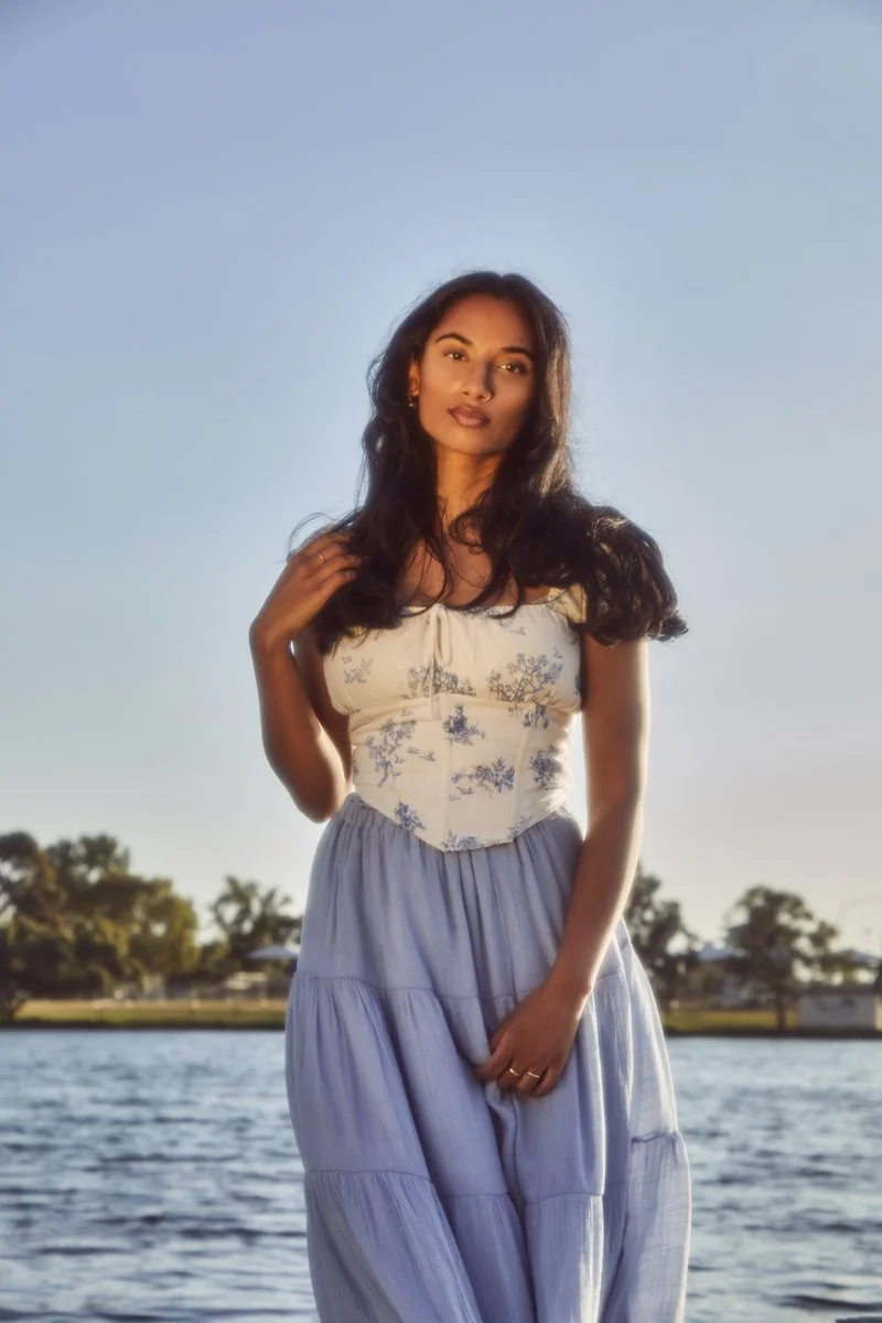 A woman standing outdoors by a body of water during sunset or sunrise. She has long dark hair, is wearing a white top with blue floral patterns and a long light blue skirt. The sky is clear and the background includes some trees and land.