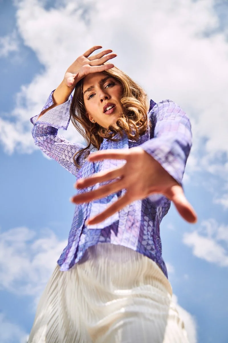 A young woman with brown wavy hair wearing a purple blouse with a geometric pattern and cream-colored pants, looking down at the camera with her hand raised above her forehead, against a partly cloudy sky.