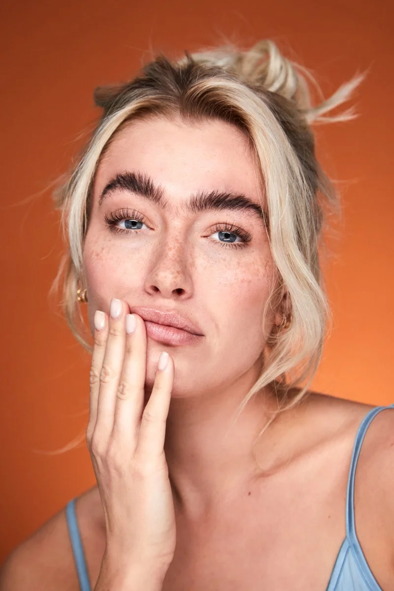 A woman with blonde hair, freckles, and blue eyes, posing with her hand near her mouth against an orange background.