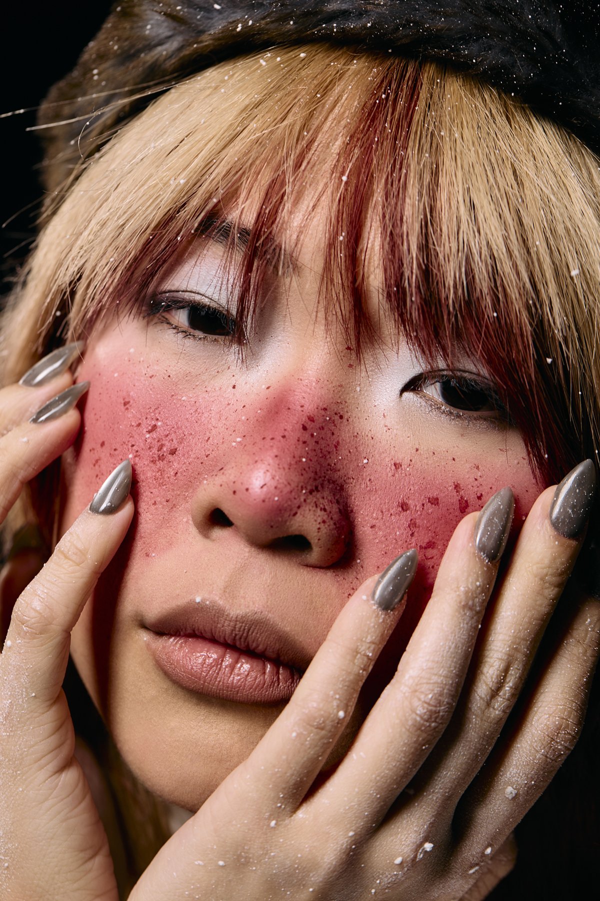 Close-up of a woman's face with splashes of red on her cheeks and nose, her eyes partially closed, and hands touching her face with long nails and powdered fingers.