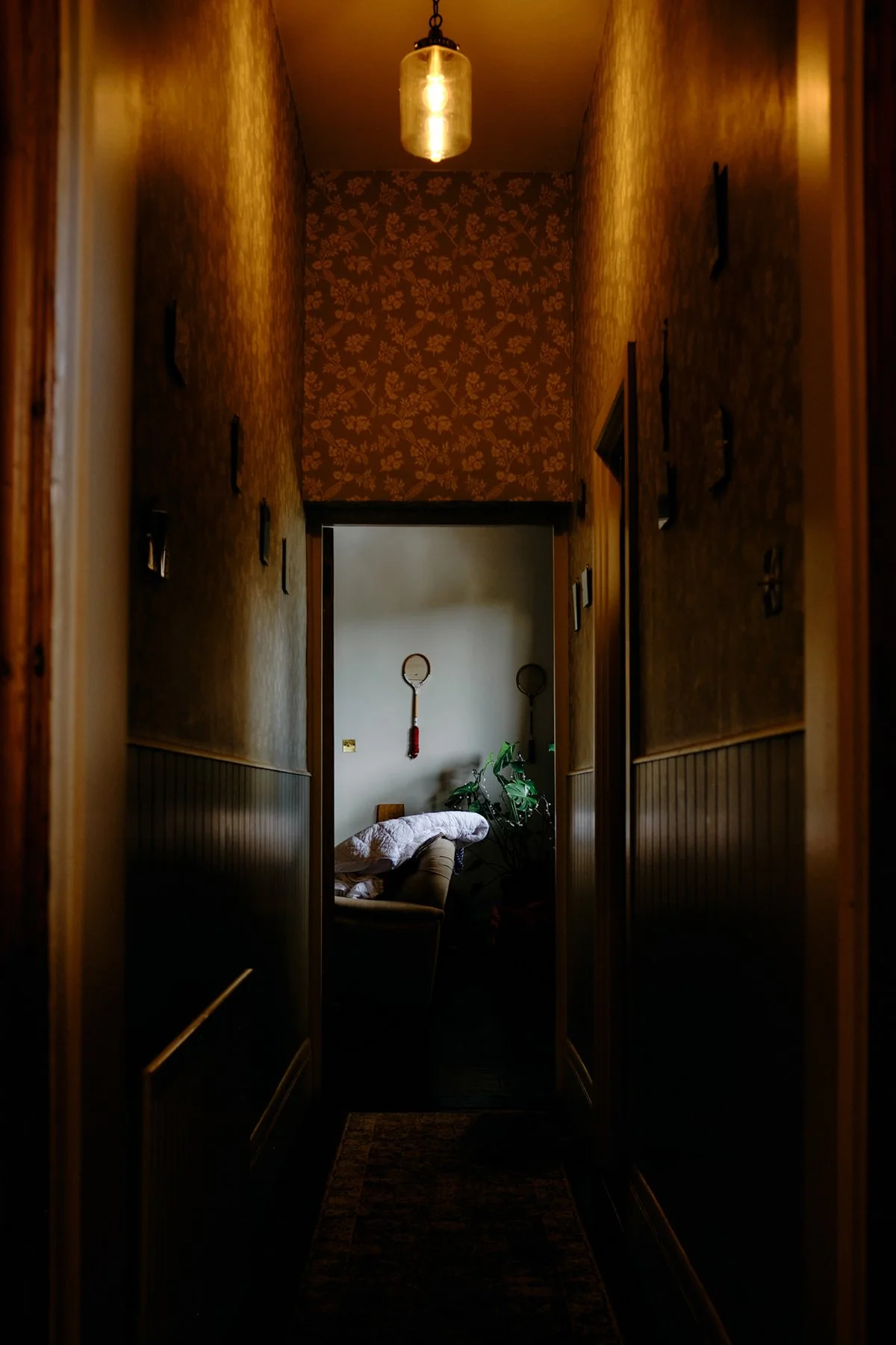 A narrow, dimly lit hallway with floral wallpaper on the far wall, leading to a room with a sofa, a potted plant, and decorative tennis rackets hanging on the wall.