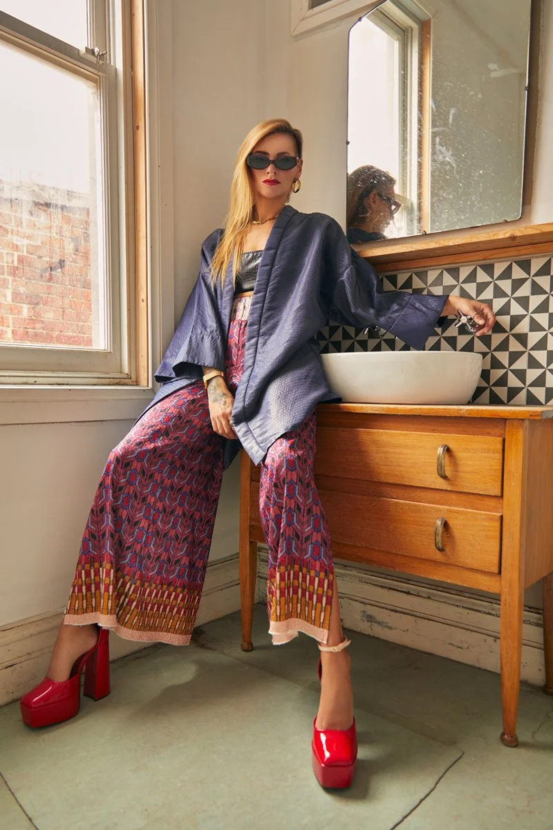 A stylish woman with blonde hair, wearing black sunglasses, red lipstick, gold jewelry, a dark blue jacket, patterned wide-leg pants, and red platform heels, is standing next to a wooden bathroom vanity with a bowl-shaped sink. She is extending her r