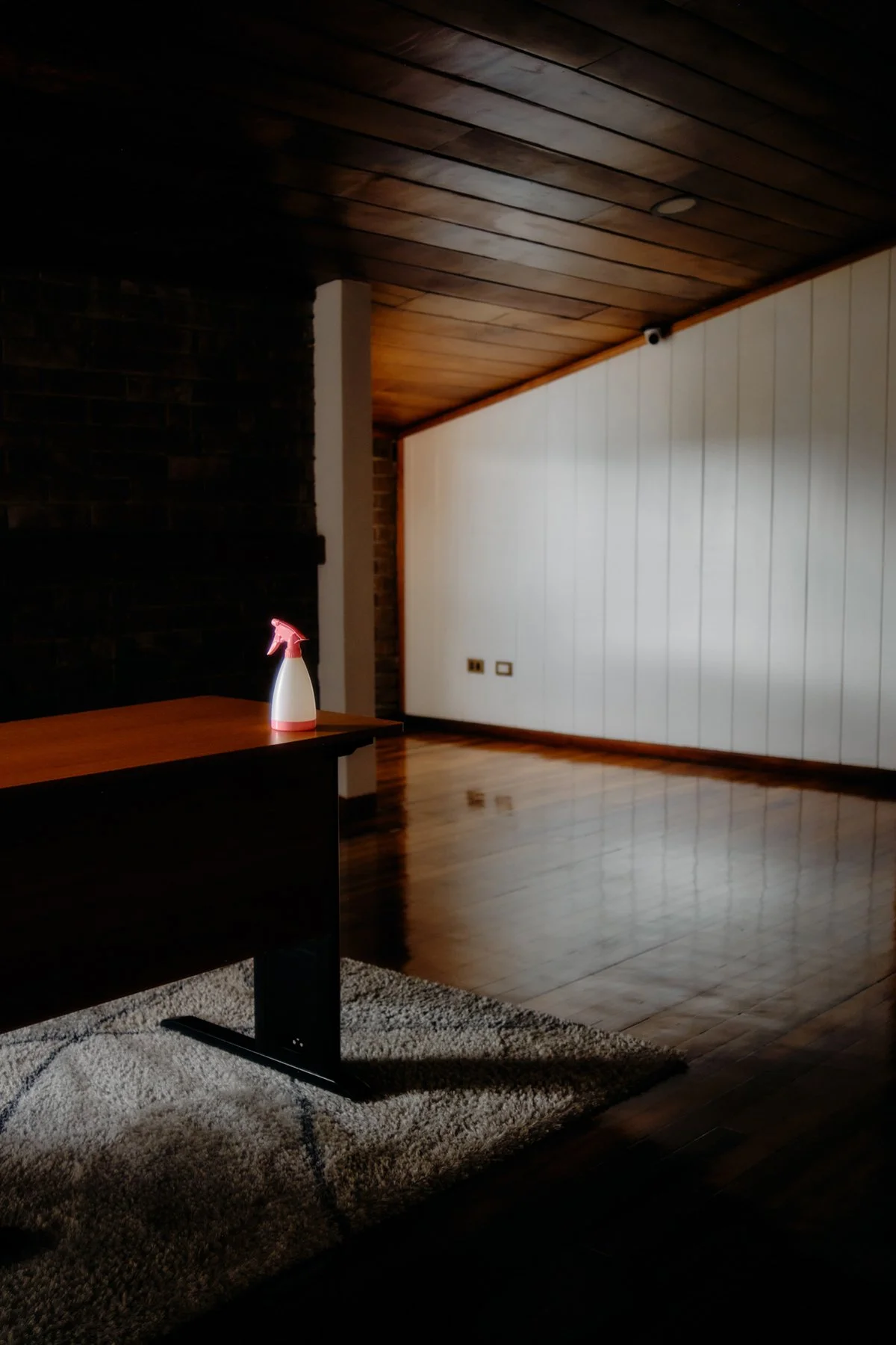 Empty room with wooden ceiling and flooring, white paneled wall, and a black brick wall. A spray bottle is placed on a wooden table on a rug.