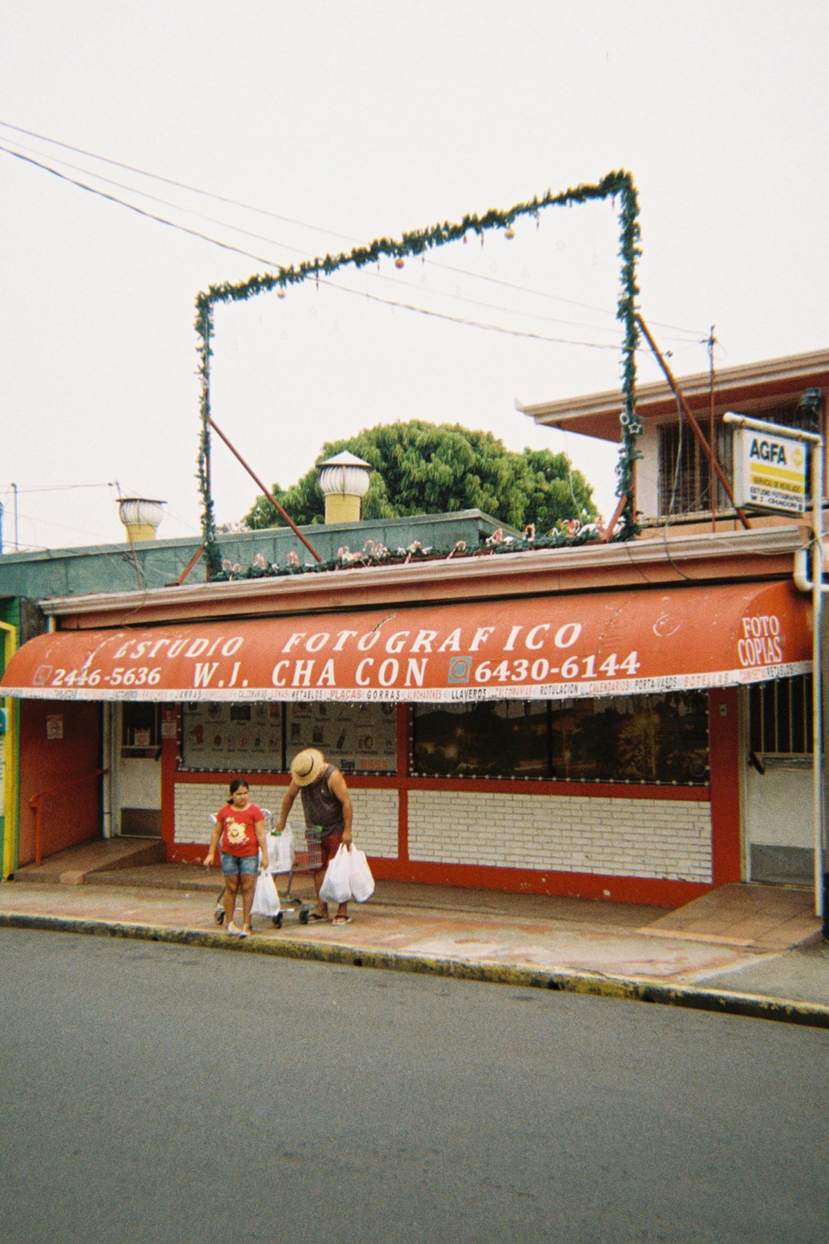 A storefront with a red awning reading 'Estudio Fotografico W. J. Cha con' and a phone number, with two people walking on the sidewalk in front, one adult and one young girl.