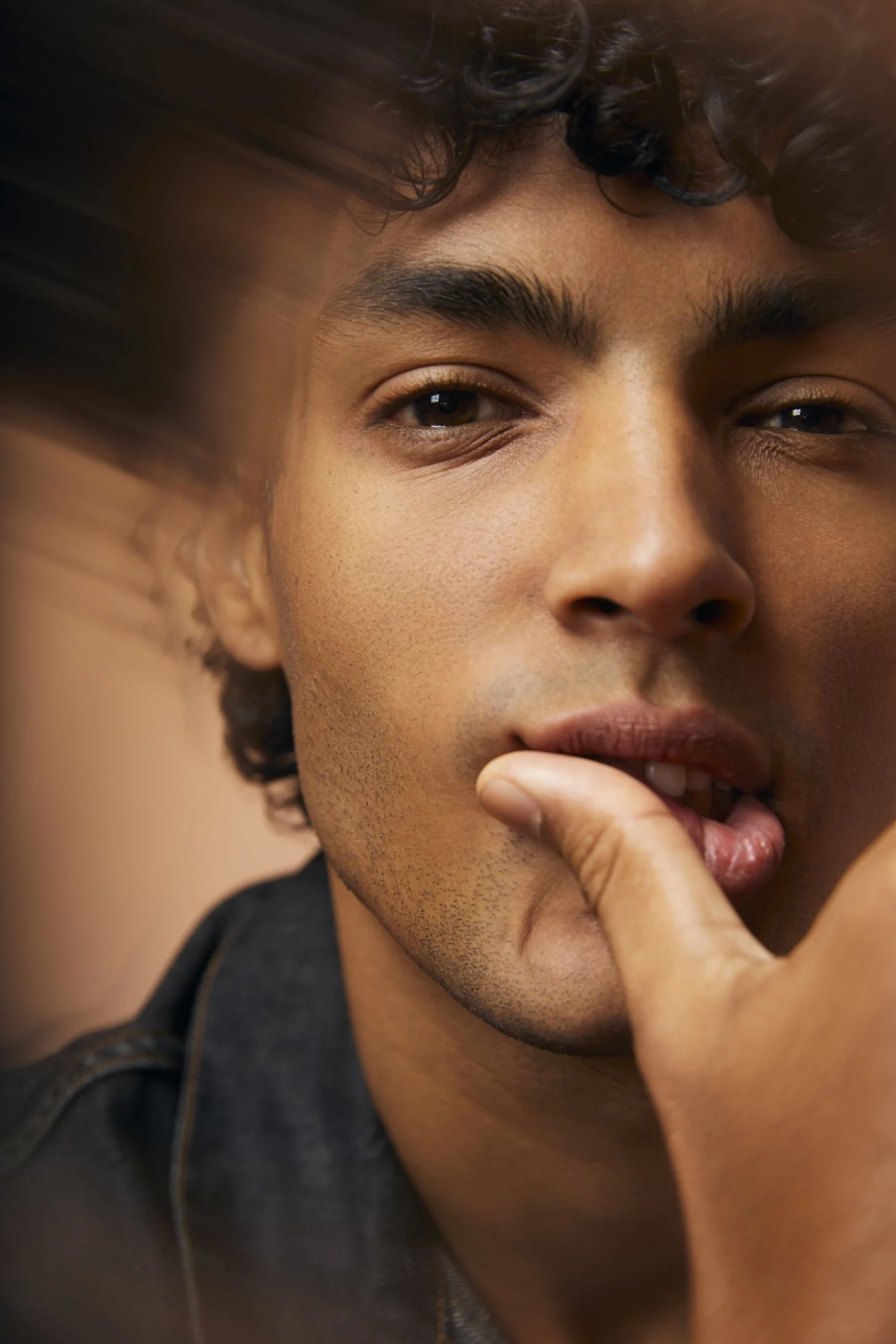 Portrait photography close-up of a young man with curly hair, raising his eyebrows slightly, with his fingers near his lips in a thoughtful pose.
