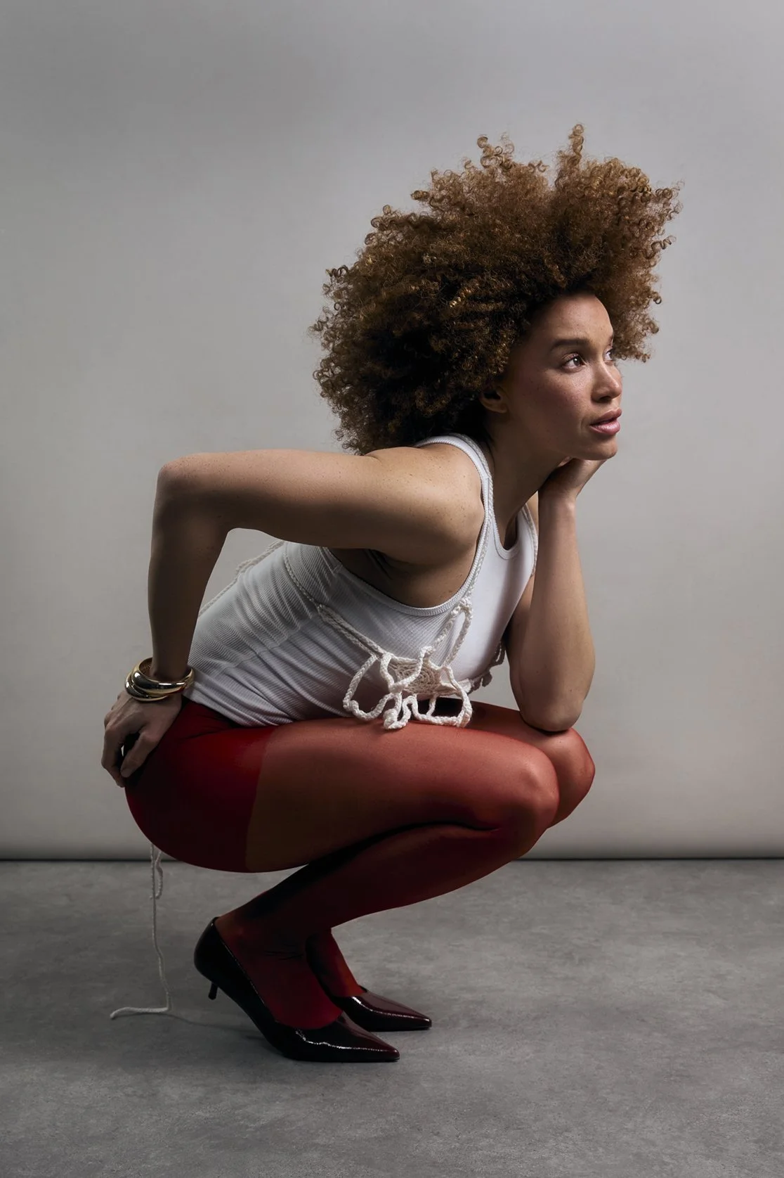 Fashion photography of a woman with curly brown hair in a squatting pose, wearing a white sleeveless top, red tights, and black heels, with a neutral background.