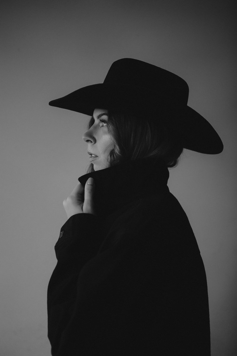 Side profile of a woman wearing a wide-brimmed hat, a high-collared coat, in black and white with a plain background.