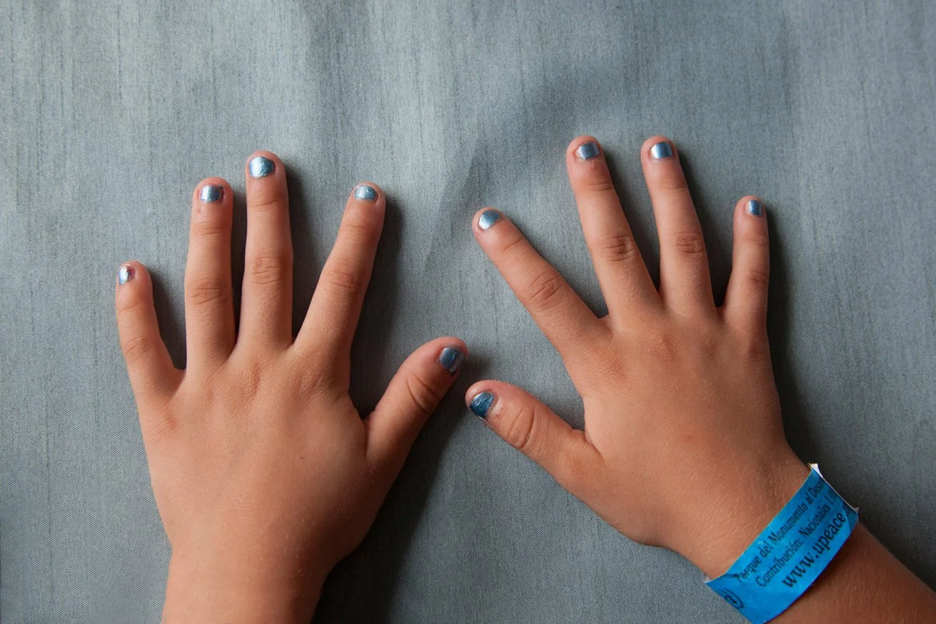 Two children’s hands with painted fingernails resting on a gray fabric surface, one of the hands has a blue wristband from a peace event.