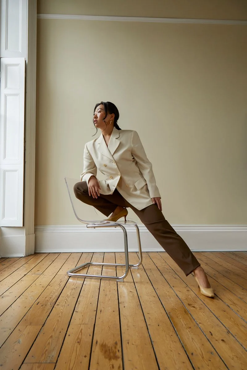 Woman in a cream blazer and brown pants striking a pose while sitting on a clear chair in a room with wooden floor and beige walls.