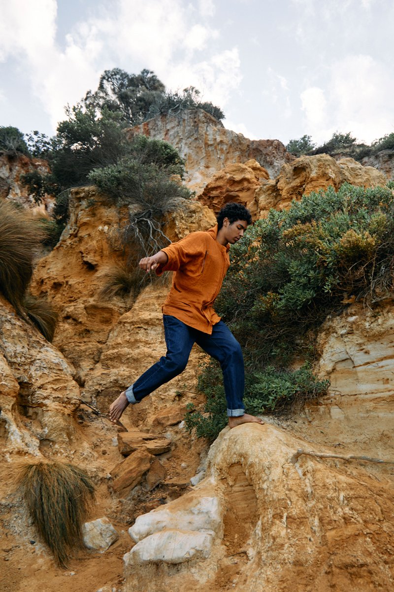 A person in an orange jacket and blue jeans barefoot, balancing on rocks while hiking in a rocky, desert-like landscape with shrubs and a cloudy sky.