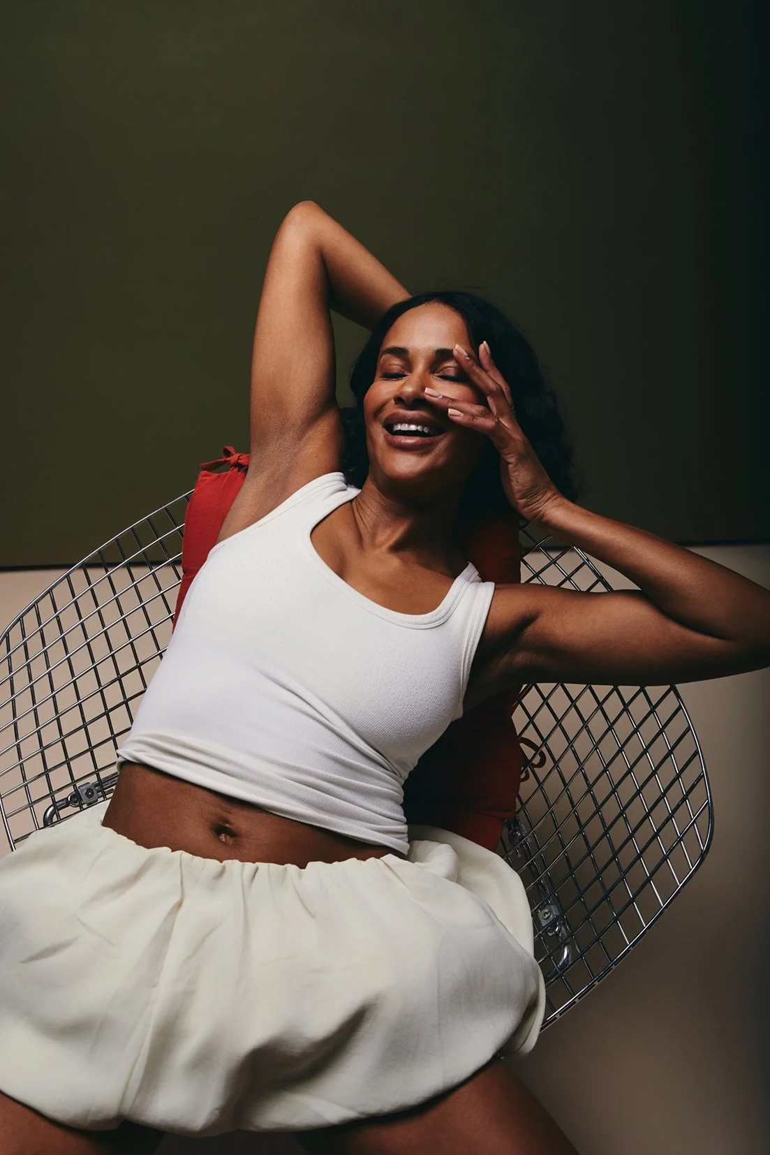 Fashion photography of a woman smiling with eyes closed, reclining on a modern wire chair, wearing a white tank top and loose beige shorts, with one arm behind her head and the other hand touching her face, against a dark green background.