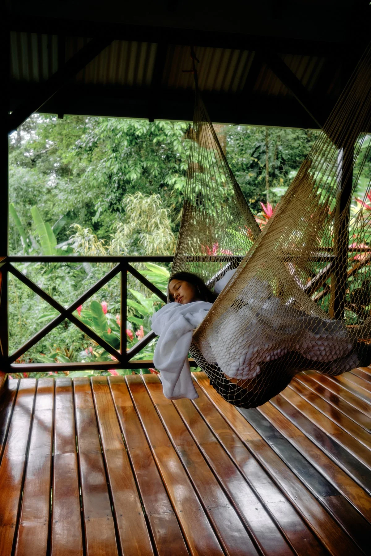 A woman resting in a hammock on a wooden deck surrounded by lush greenery and colorful flowering plants.