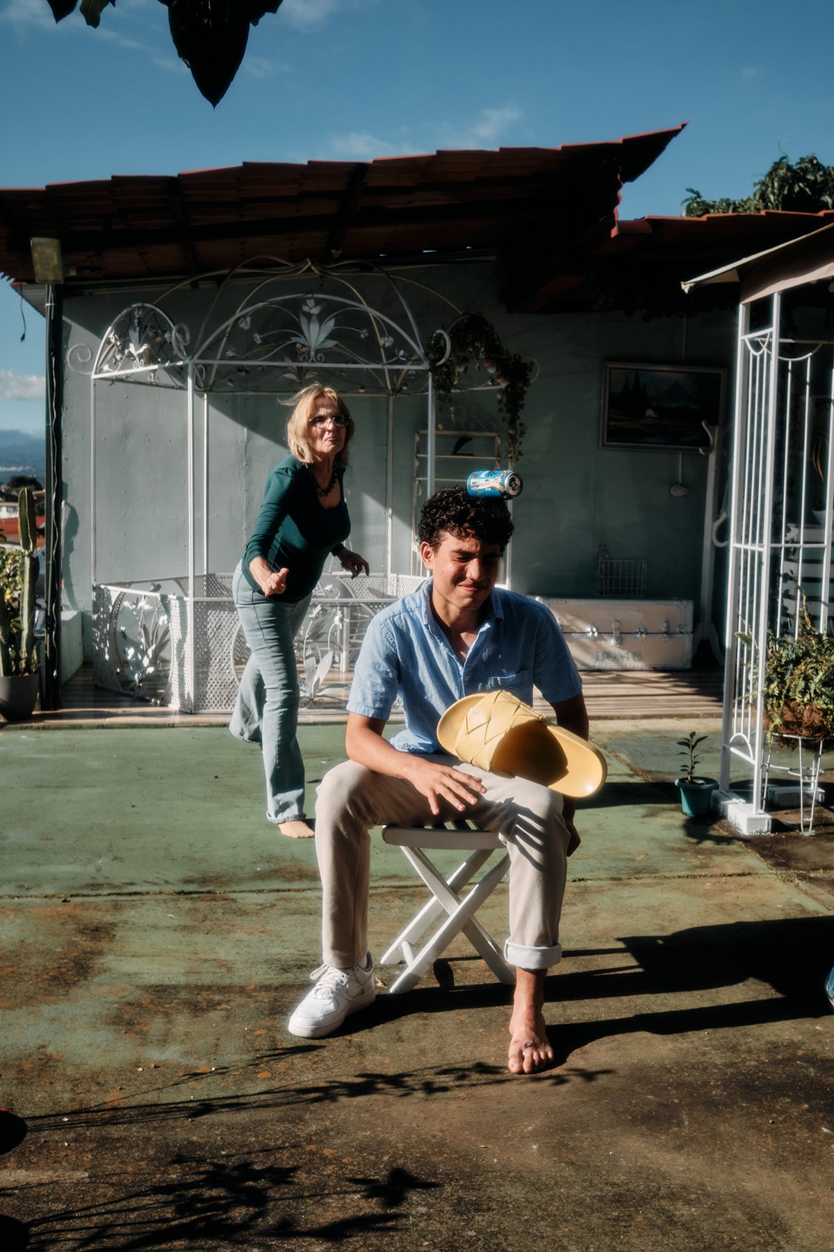 A woman throws a show towards a young man sitting outside on a folding chair, trying to knock a can balanced on his head. The scene is outdoors, with a house and potted plants in the background, and the sunlight is casting distinct shadows.