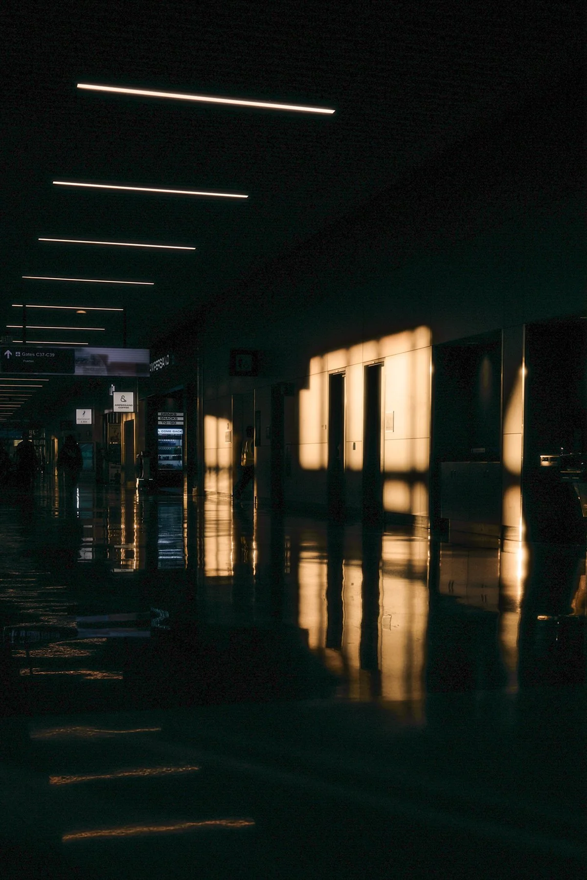 An airport terminal with sunlight streaming through windows, casting reflections and shadows on the floor, with a few travelers visible in the distance.