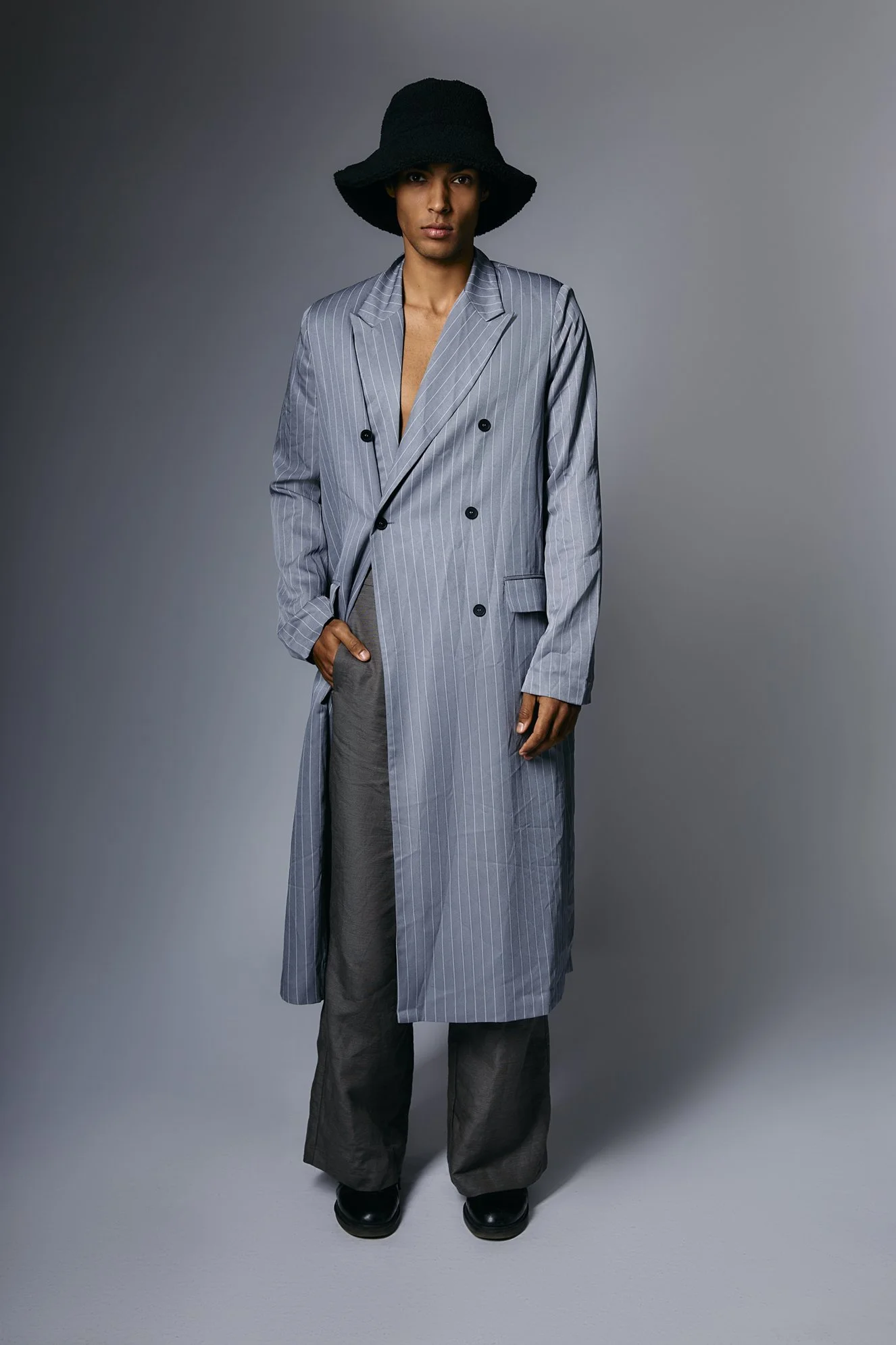 Fashion photography of a person wearing a black floppy hat, a gray pinstripe double-breasted coat, gray pants, and black shoes, standing against a neutral background.