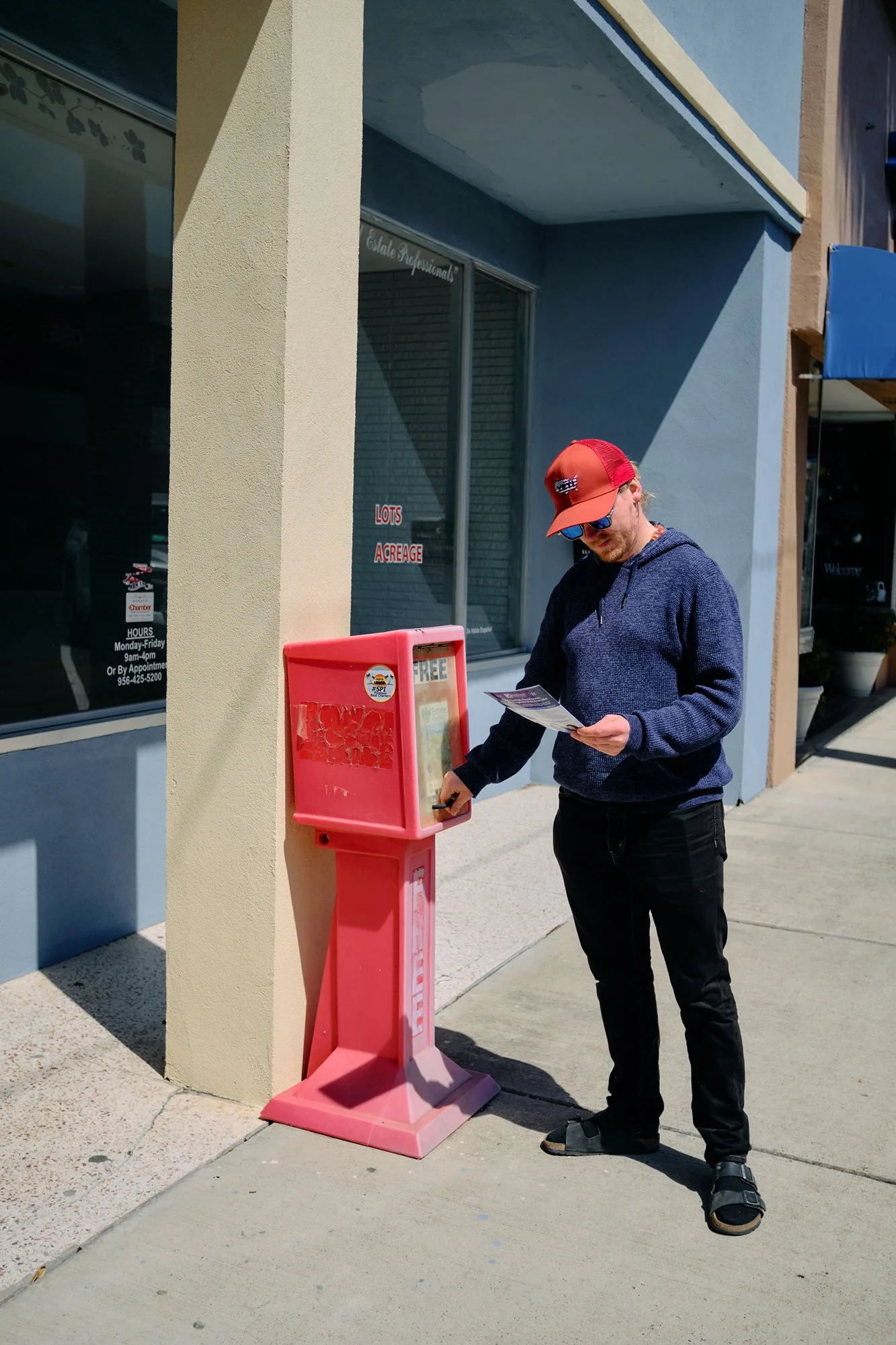 Travel photography of a man wearing a red cap, sunglasses, and a dark blue hoodie stands outside a storefront, reading a flyer or pamphlet while holding a pen, next to a pink mailbox on the sidewalk.
