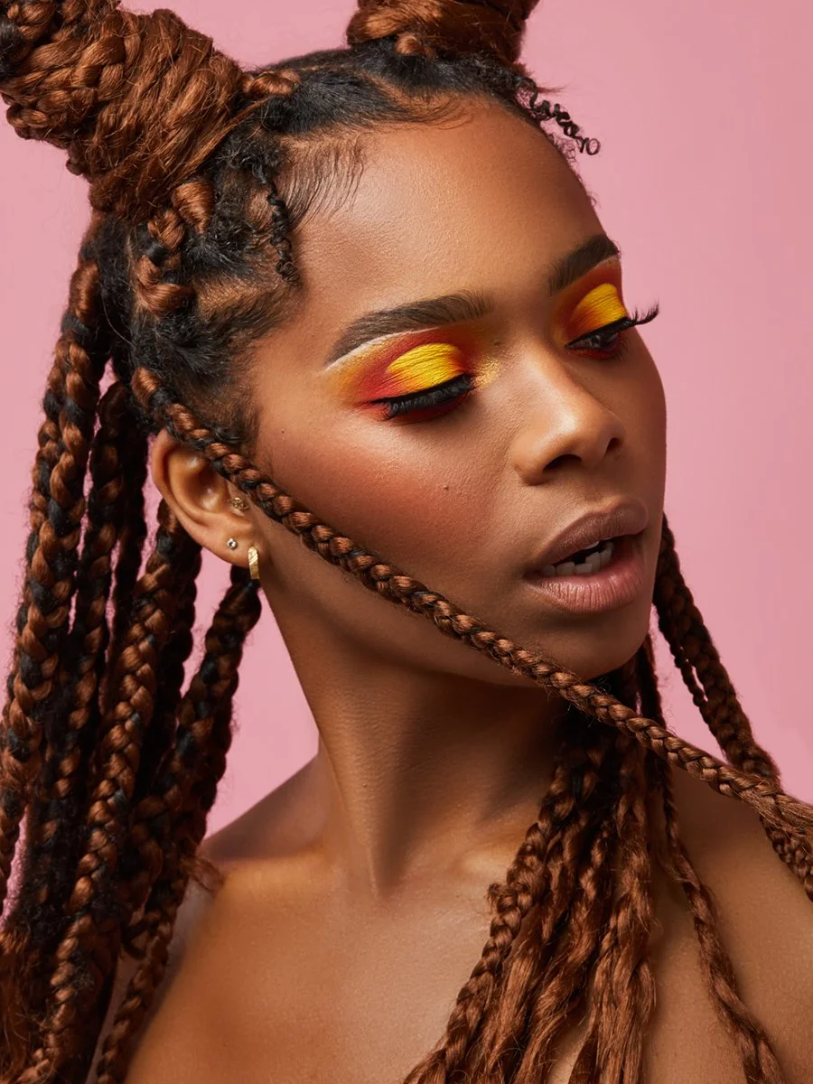 A woman with long, braided, brown hair styled in buns and loose braids, wearing vibrant yellow and orange eyeshadow with bold eyeliner, against a pink background.