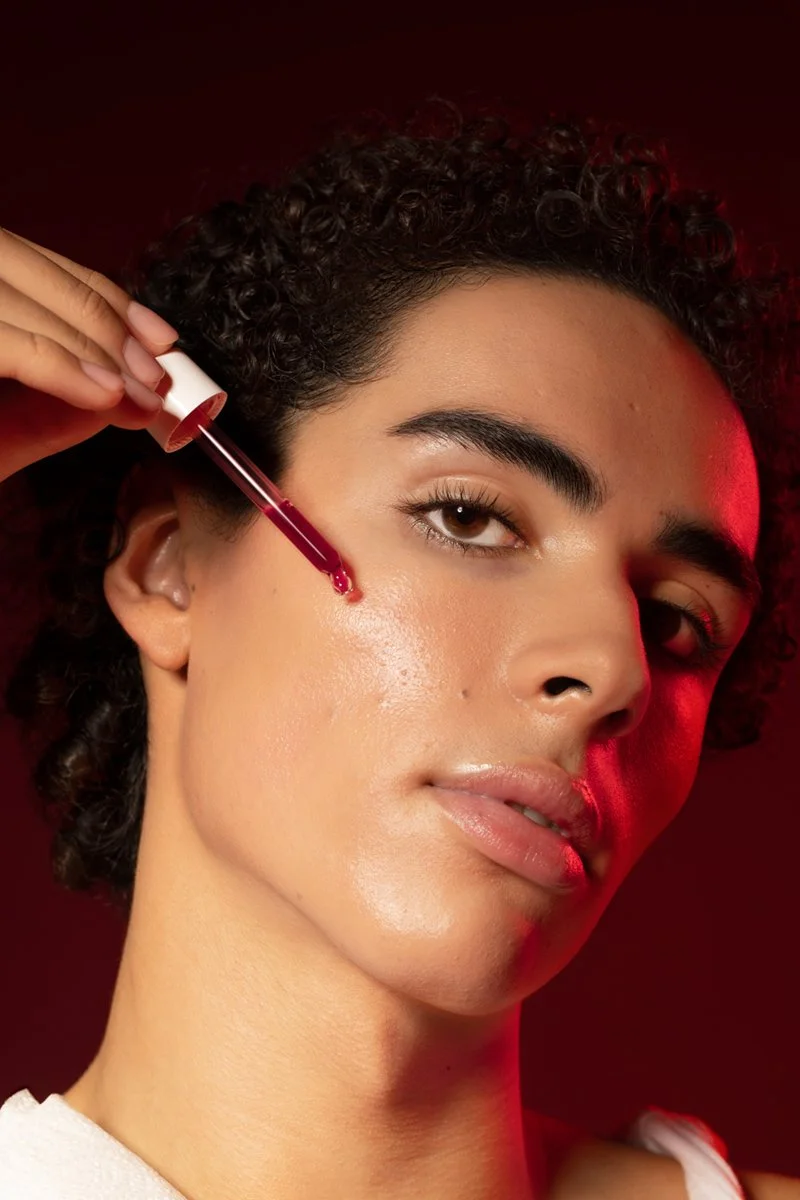 A woman with curly hair applying facial serum with a dropper on her cheek, standing against a dark red background.