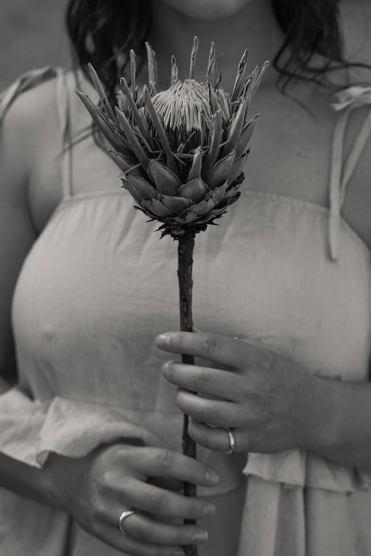 A woman in a sleeveless top is holding a large artichoke flower on a stick. The image is in black and white.