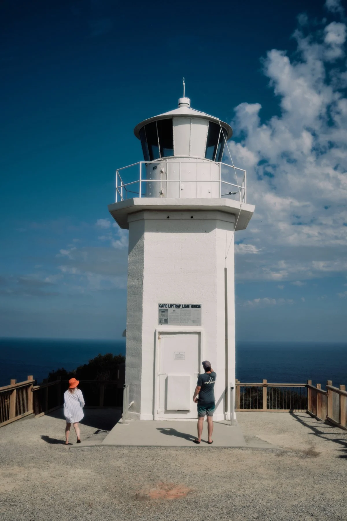 A white lighthouse labeled Cape Liptrap Lighthouse under a partly cloudy sky with two people standing near its base, overlooking the ocean.