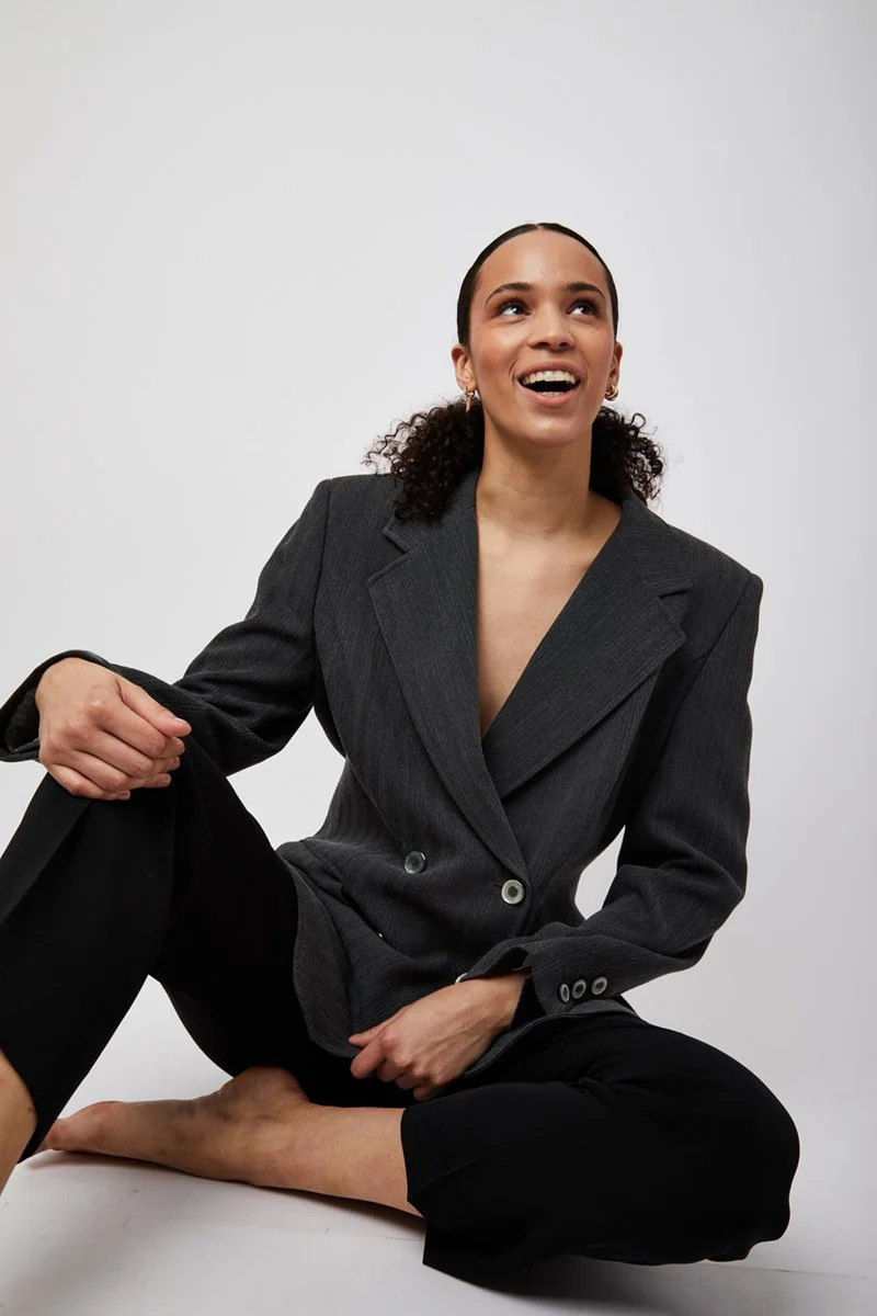 A woman sitting on the floor in a gray blazer and black pants, smiling and looking up, with plain white background.