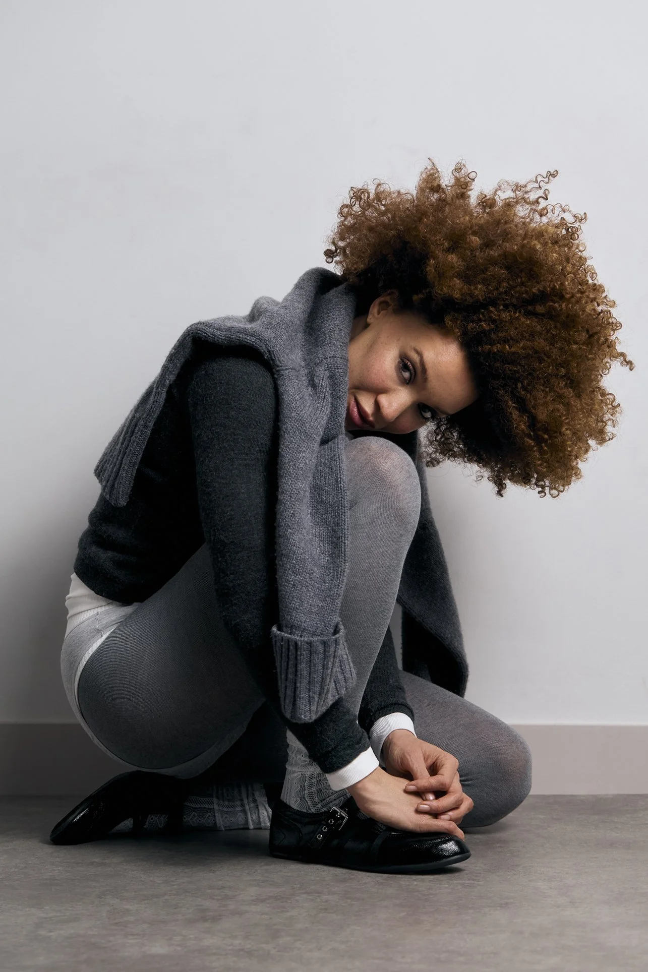 Portrait photography of a young woman with curly hair sitting on the floor, wearing a gray vest over black top, gray pants, and black shoes, against a plain light background.