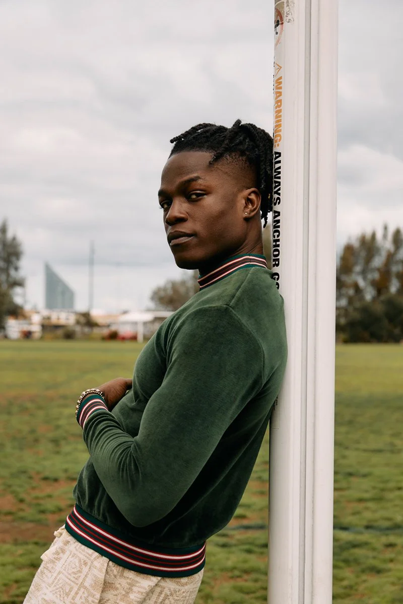 A young man with dark skin and dreadlocks, wearing a green velour jacket, stands with arms crossed, leaning against a white pole in an outdoor park with cloudy sky and buildings in the background.