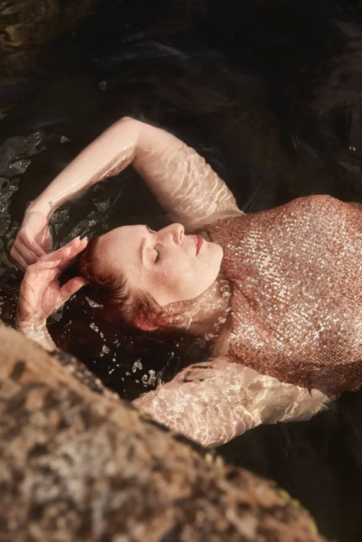Woman with red hair, closed eyes, wearing a sparkly pink top, floating in water with her arm resting on her forehead.
