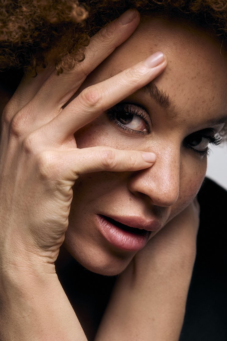 Close-up of a woman with curly hair, one eye visible, partially covering her face with her hand, showing freckles, makeup, and a playful expression for a beauty campaign.