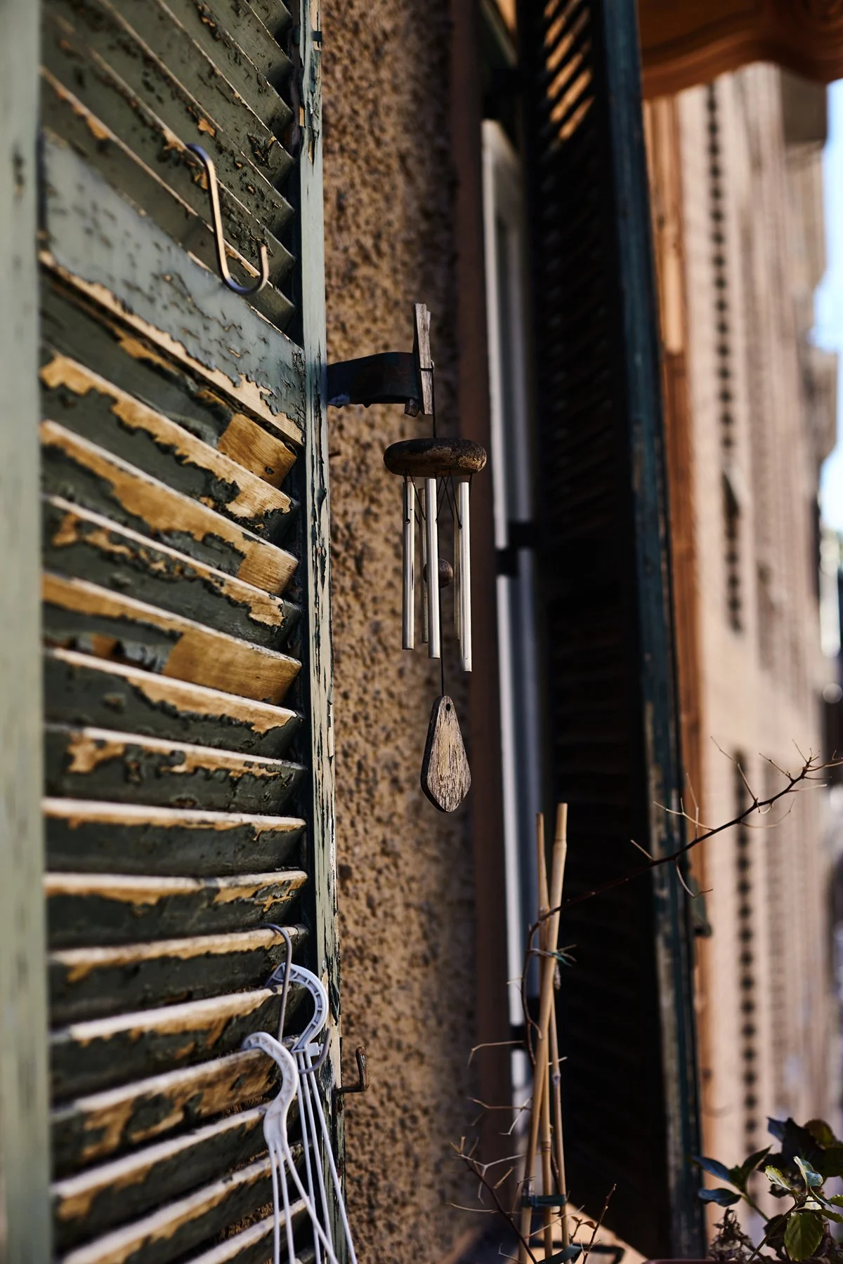 Close-up of a weathered green window shutter with peeling paint, next to a textured stucco wall. Hanging on the wall are a rustic wind chime with metal tubes and a wooden paddle, and several plastic hangers. Another window with open dark shutters is 