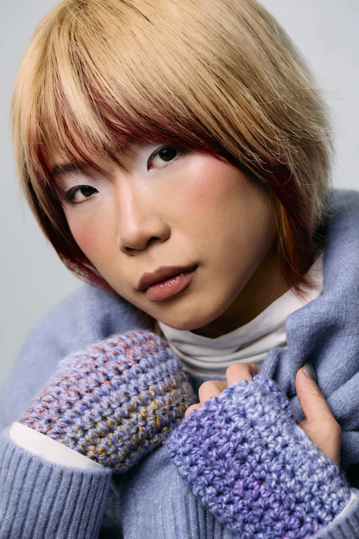 A person with short, layered, reddish-brown hair looking at the camera with a neutral expression, wearing a light blue sweater with multicolored knitted gloves and a white shirt underneath.