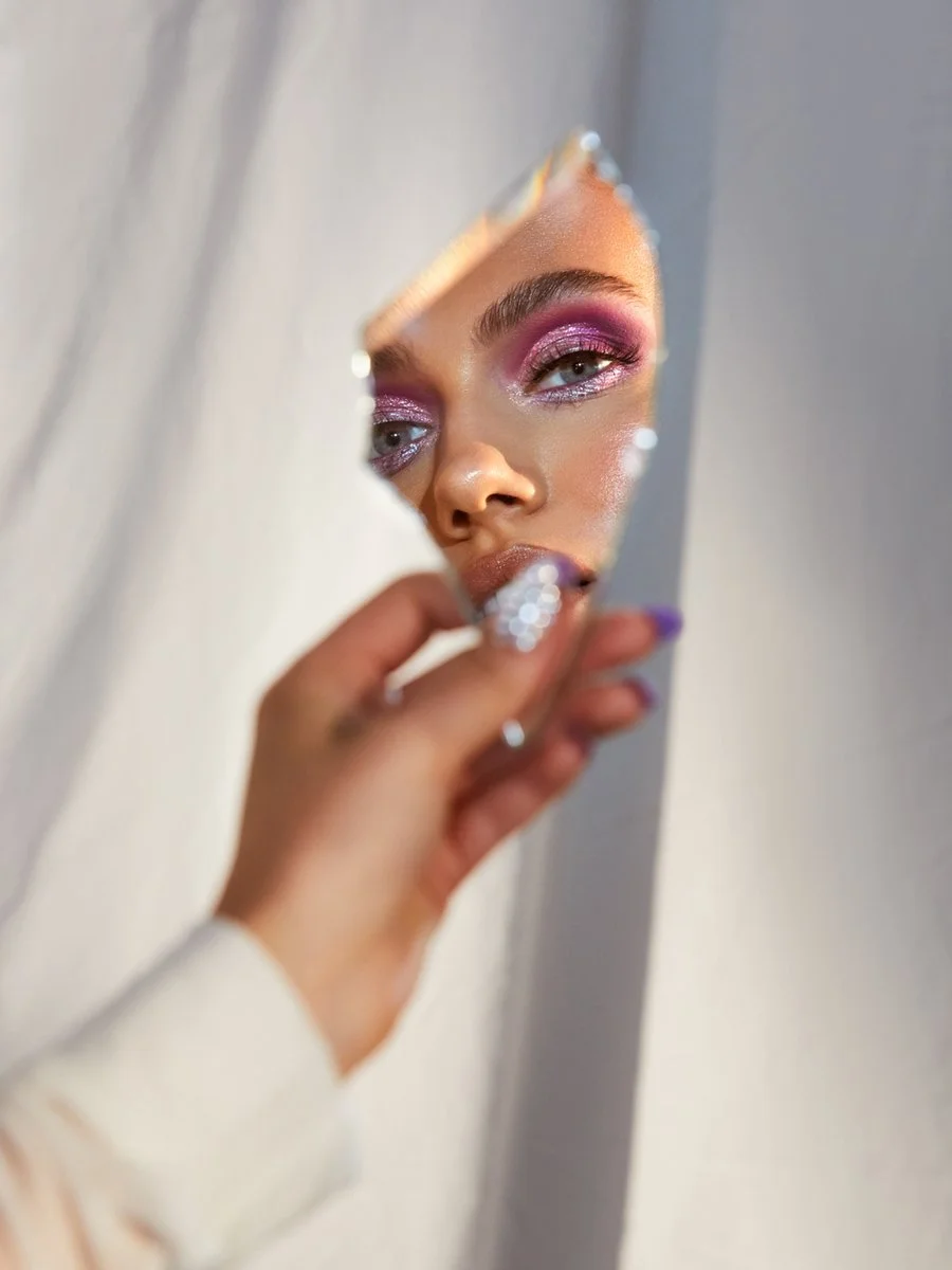Close-up of a woman applying makeup while looking in a small mirror, with pink eyeshadow and glossy lips.