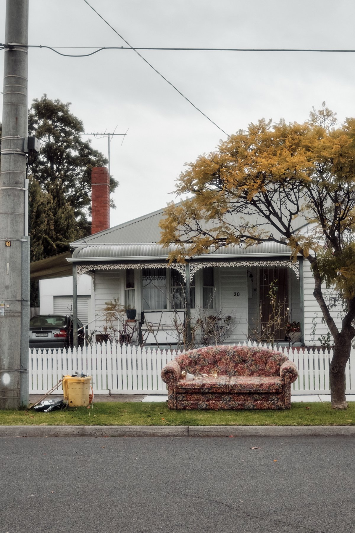 A vintage floral upholstered sofa on the sidewalk in front of a white picket fence, with a house, tree, and power pole in the background.