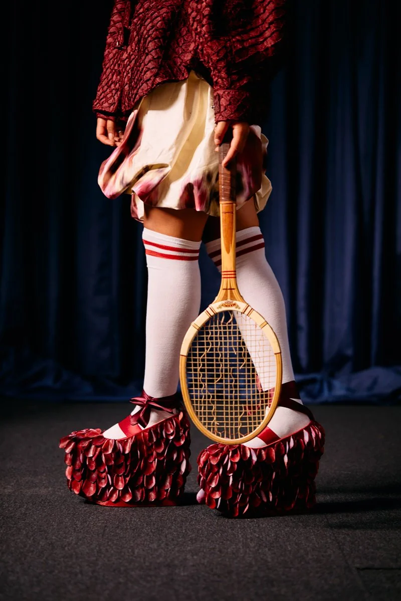 Person wearing flamboyant tennis shoes with red petal-like details, white knee-high socks with red stripes, a dress with a patterned hem, a textured red jacket, and holding a vintage wooden tennis racket.