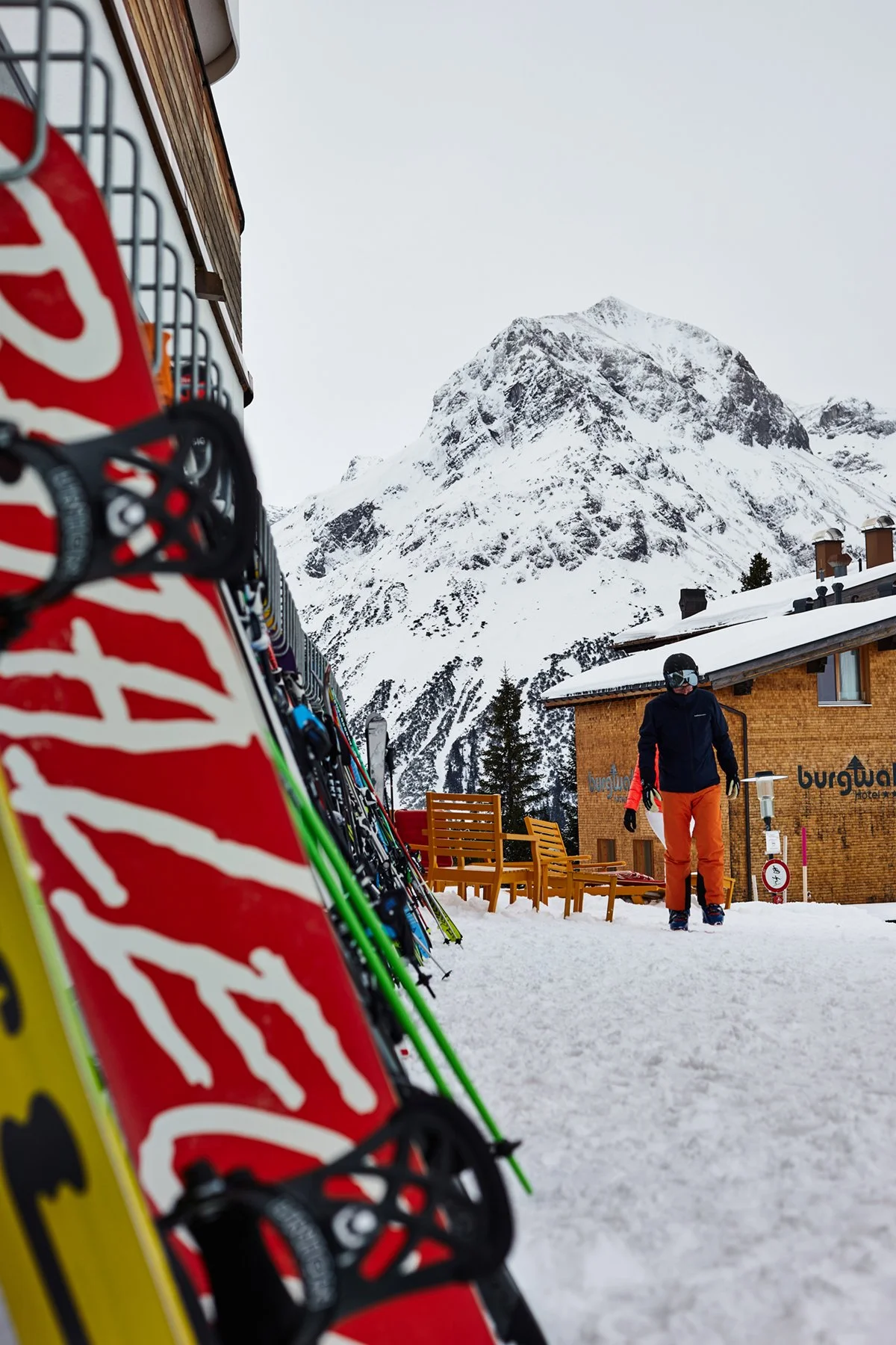 Ski equipment leaning against a wooden railing with snow-covered mountains in the background. A person dressed in ski gear and orange pants walking near a wooden building.