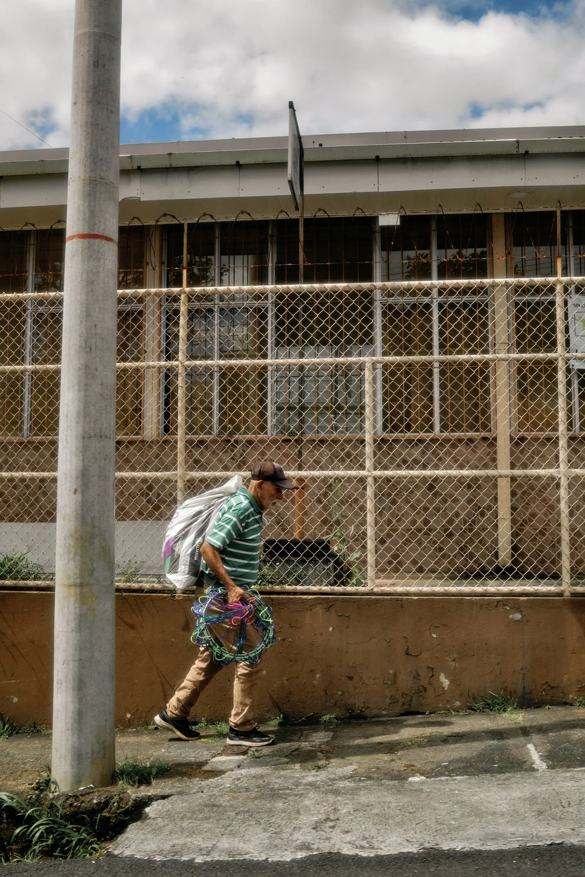 A man wearing a striped green and white shirt, beige pants, and a black cap walking along the sidewalk while carrying a bundle of colorful wire or string. He has a backpack on his back and is walking past a chain-link fence in front of an older build