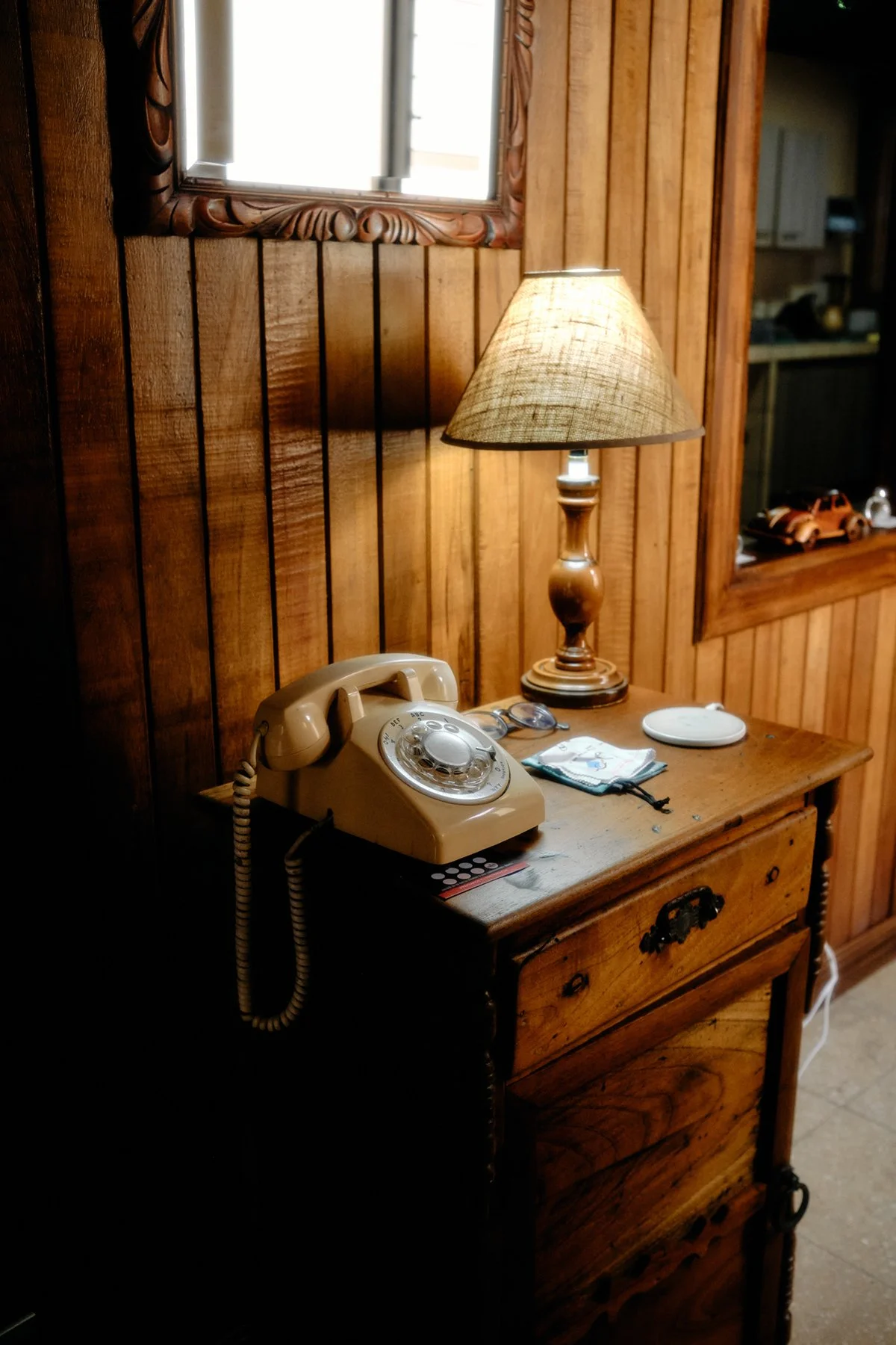 A wooden nightstand with a vintage rotary phone, a pair of eyeglasses, a small white dish, a cloth, and a lamp with a beige shade, set against a wooden-paneled wall with a mirror reflecting a window.