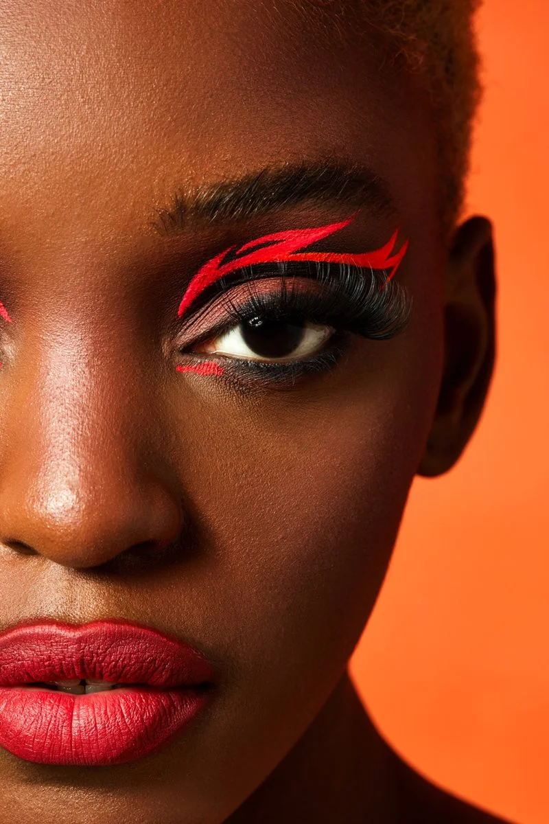 Close-up of a woman with bold red and black eye makeup, including dramatic red graphic eyeliner with flame-like designs, and matching red lipstick, against an orange background.