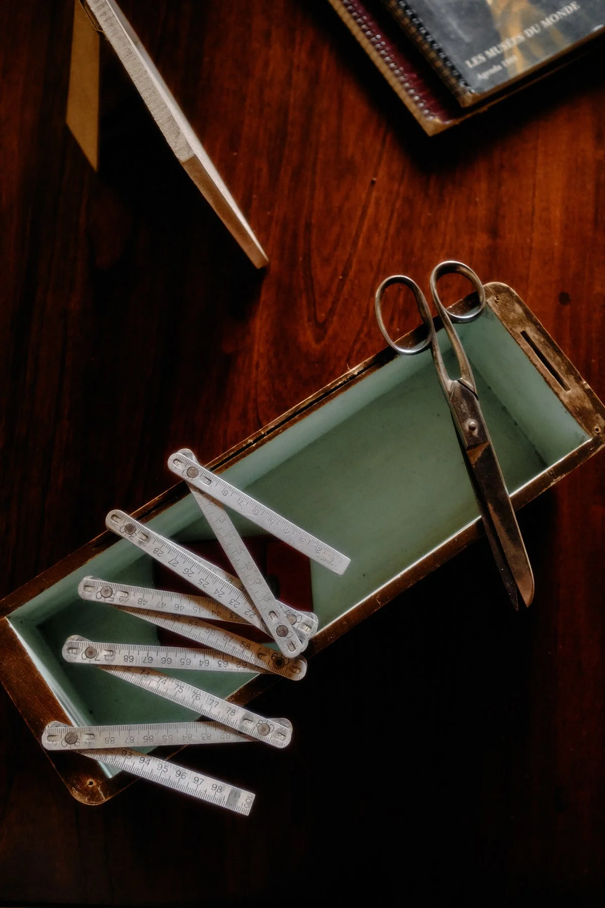 A wooden table with a light green rectangular box holding various aluminum rulers, some opened, a pair of scissors, and a partially visible book on the top right.