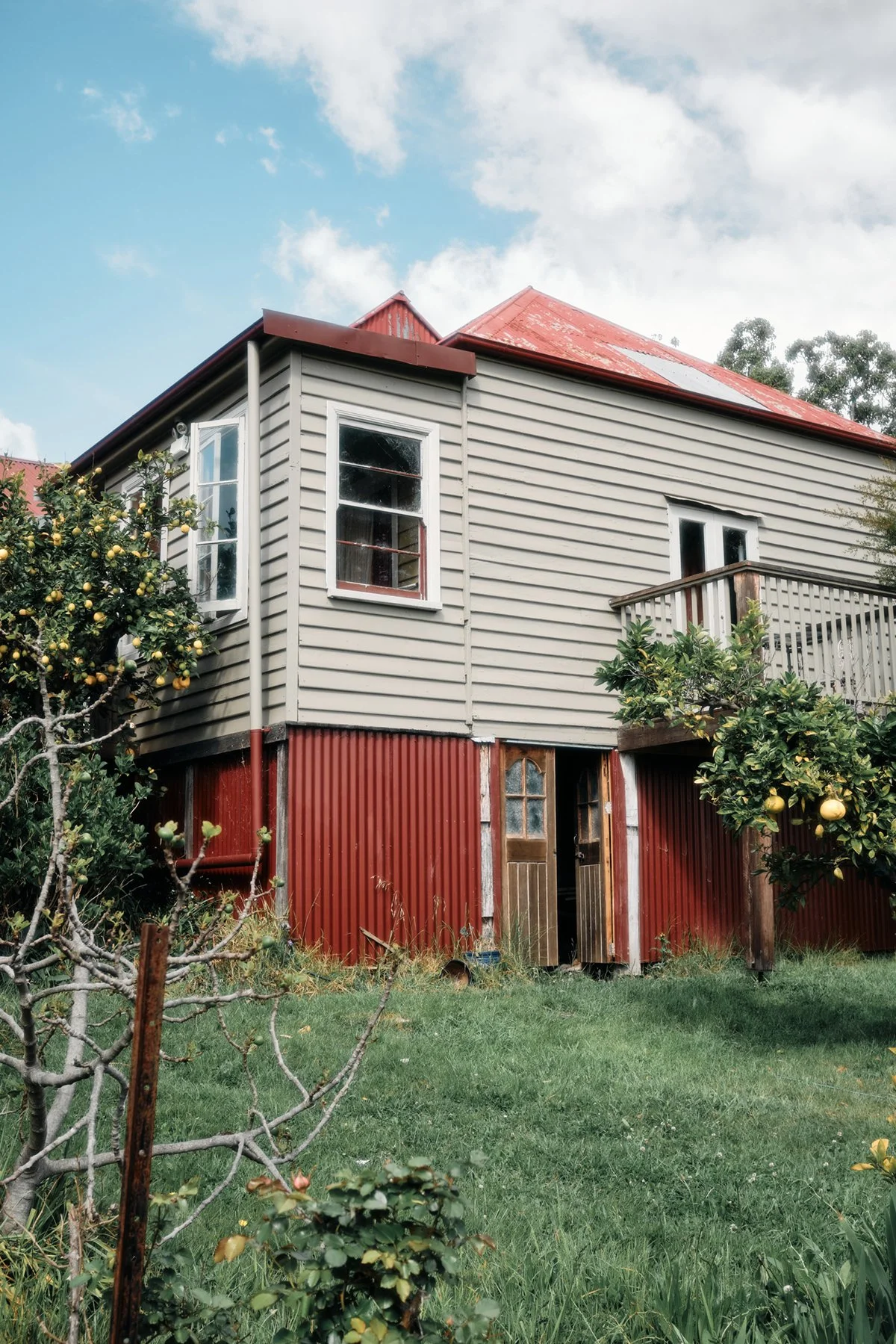 A two-story house with beige siding on the upper level and red corrugated metal on the lower level, with a balcony on the upper level and a partially open door on the lower level, surrounded by trees and greenery.