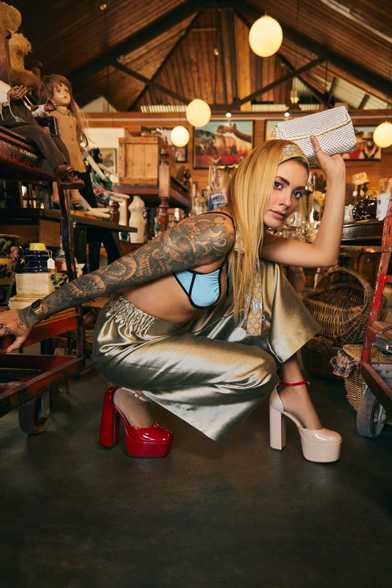 A woman with blonde hair, tattoos on her arms, wearing a blue top, metallic wide-leg pants, and two-tone high heels, crouches on the floor holding a white clutch bag in an antique store filled with various collectibles, dolls, and artwork.