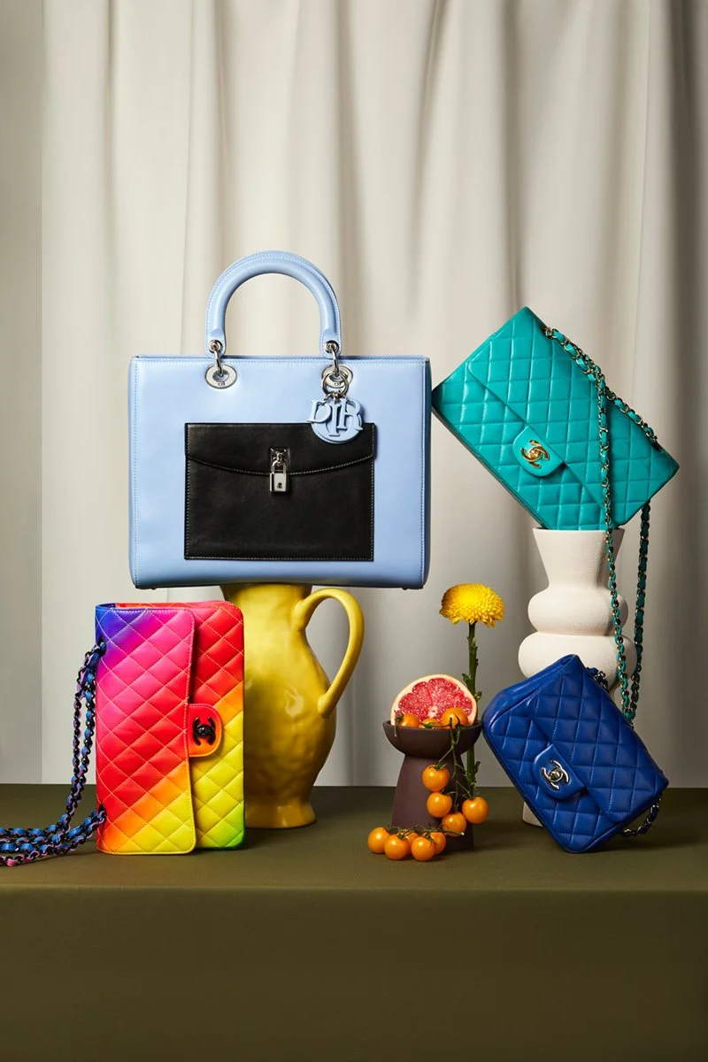 Colorful designer handbags and small decorative items arranged on a table, with curtain background.
