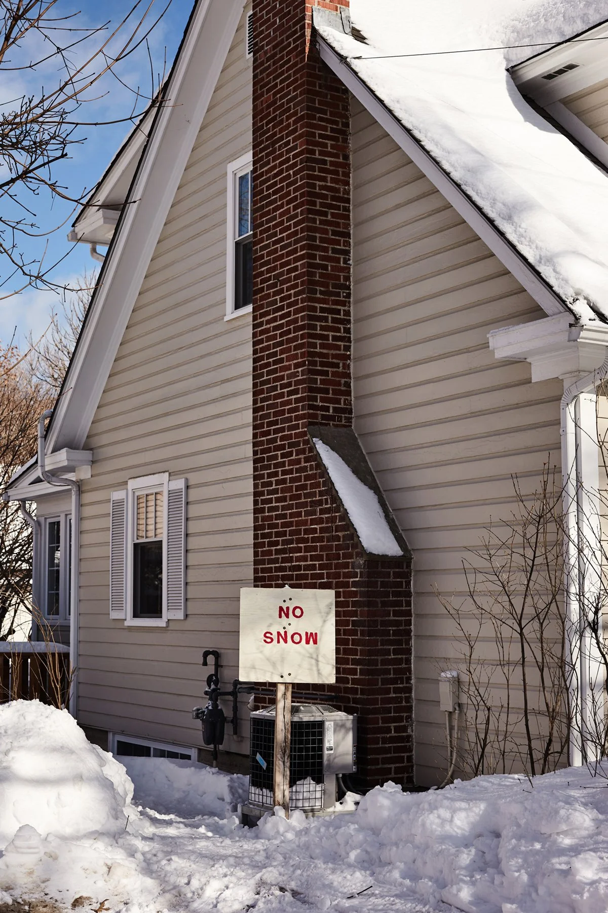 A house with beige siding and a red brick chimney, snow on the roof and ground, and a sign that says 'NO SNOW' in red text.