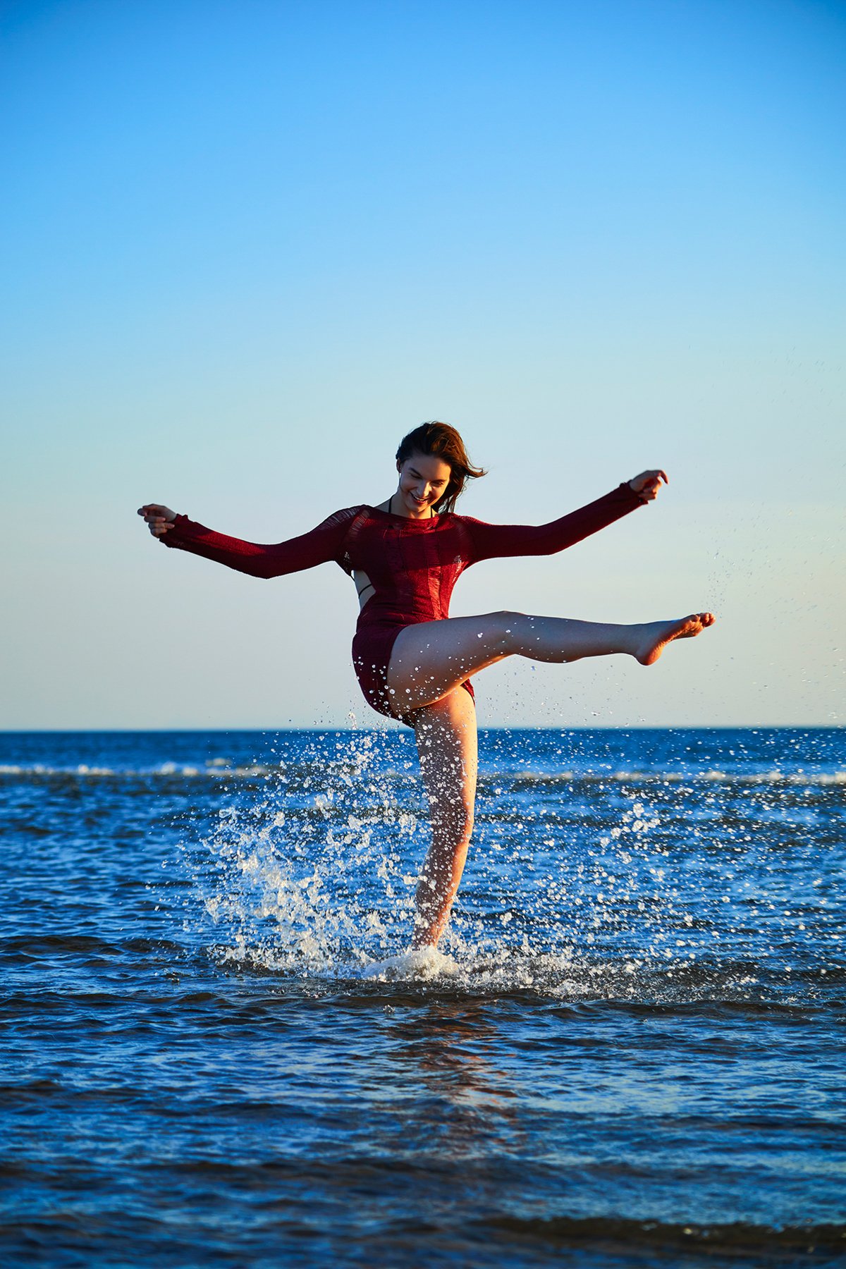 A woman in a red dress splashing water and kicking her leg high in the air at the beach during sunset.