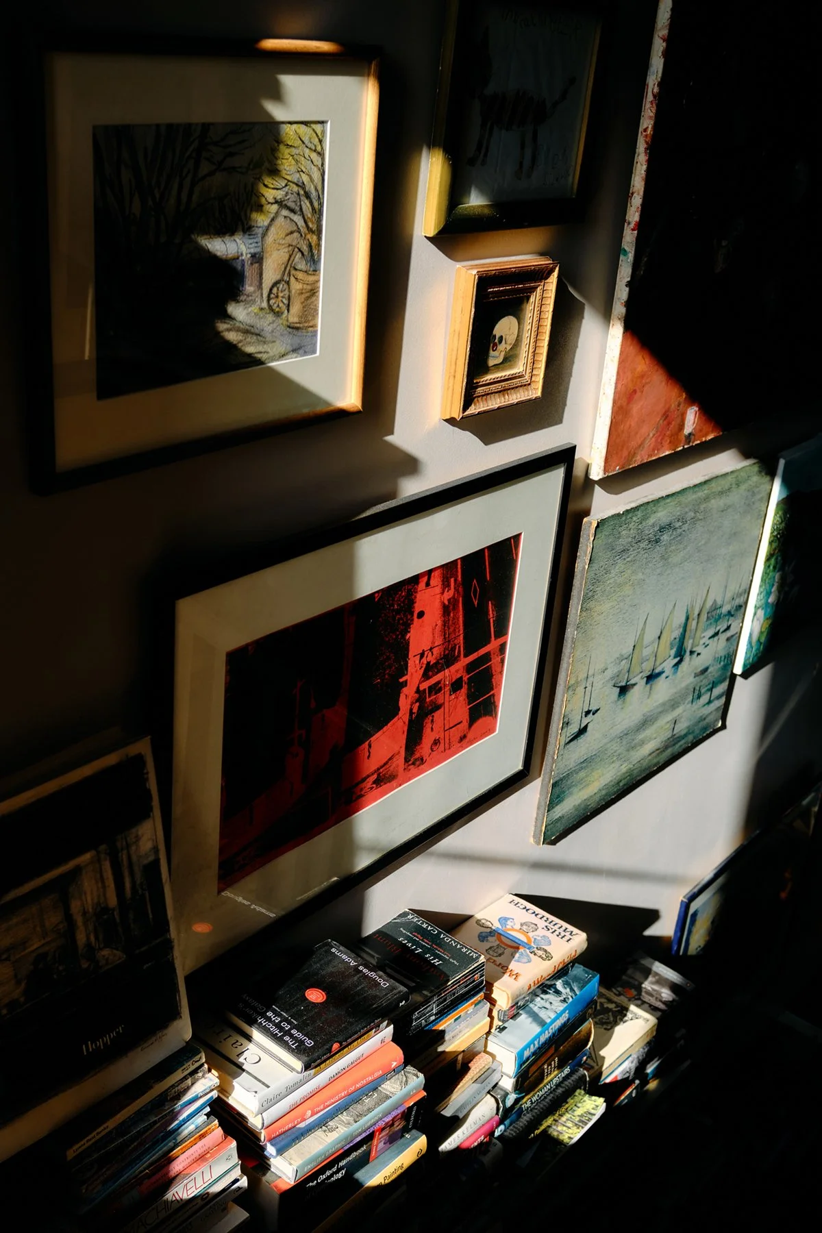A collection of framed artworks on a wall, including landscape scenes, a skull, and abstract images, with sunlight casting shadows. Below the artwork, there is a shelf filled with a stack of books.